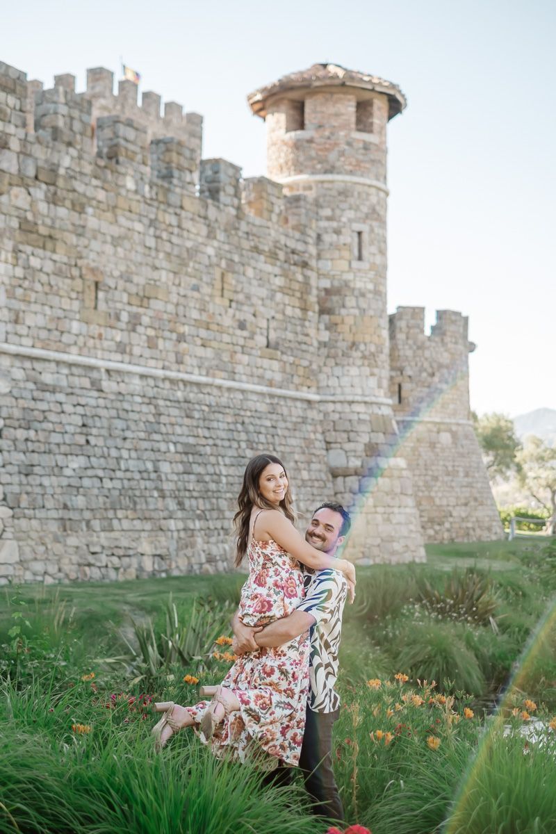 Couple embracing and laughing in front of a stone castle surrounded by lush greenery and flowers on a sunny day.