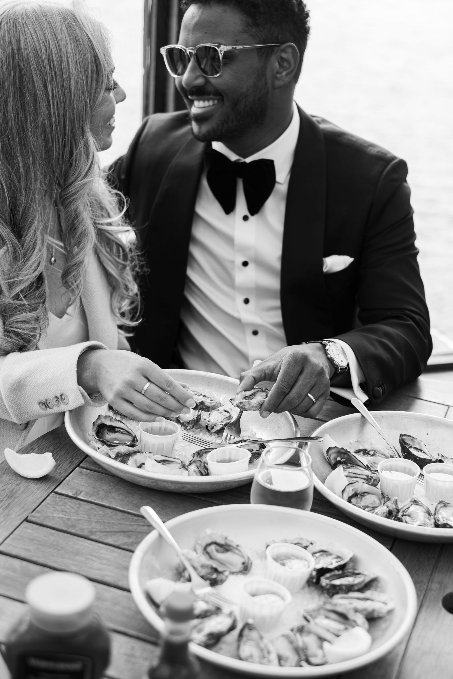 Black and white photo of a smiling couple in formal wear enjoying oysters at their wedding reception.