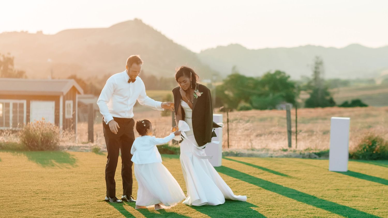A bride and groom interact with a flower girl on a lush green lawn at golden hour with mountains in the background.