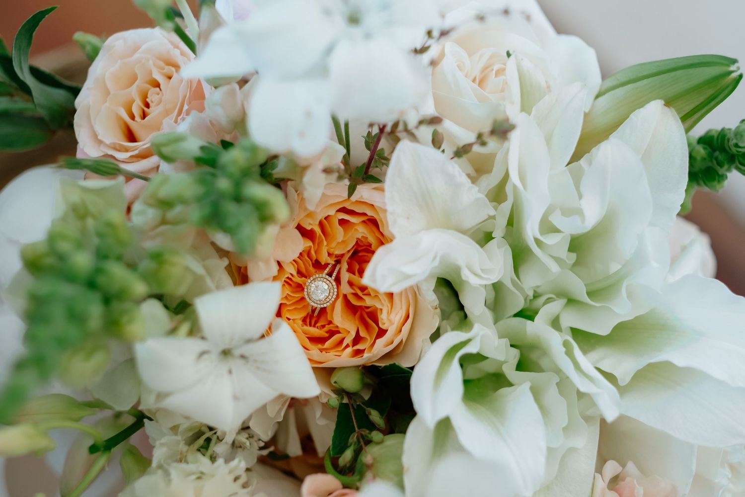 Elegant wedding bouquet featuring peach garden roses, white orchids, and green accents with a diamond ring nestled among the blooms.