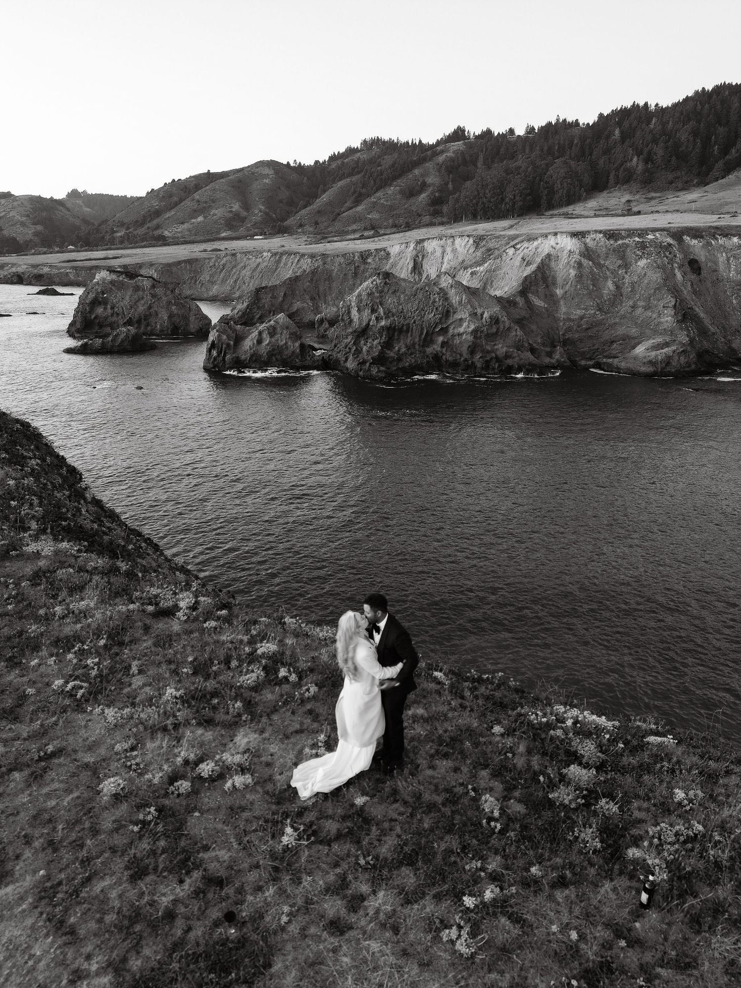 Black and white aerial photo of bride and groom kissing on coastal cliffside with ocean and rocks below.