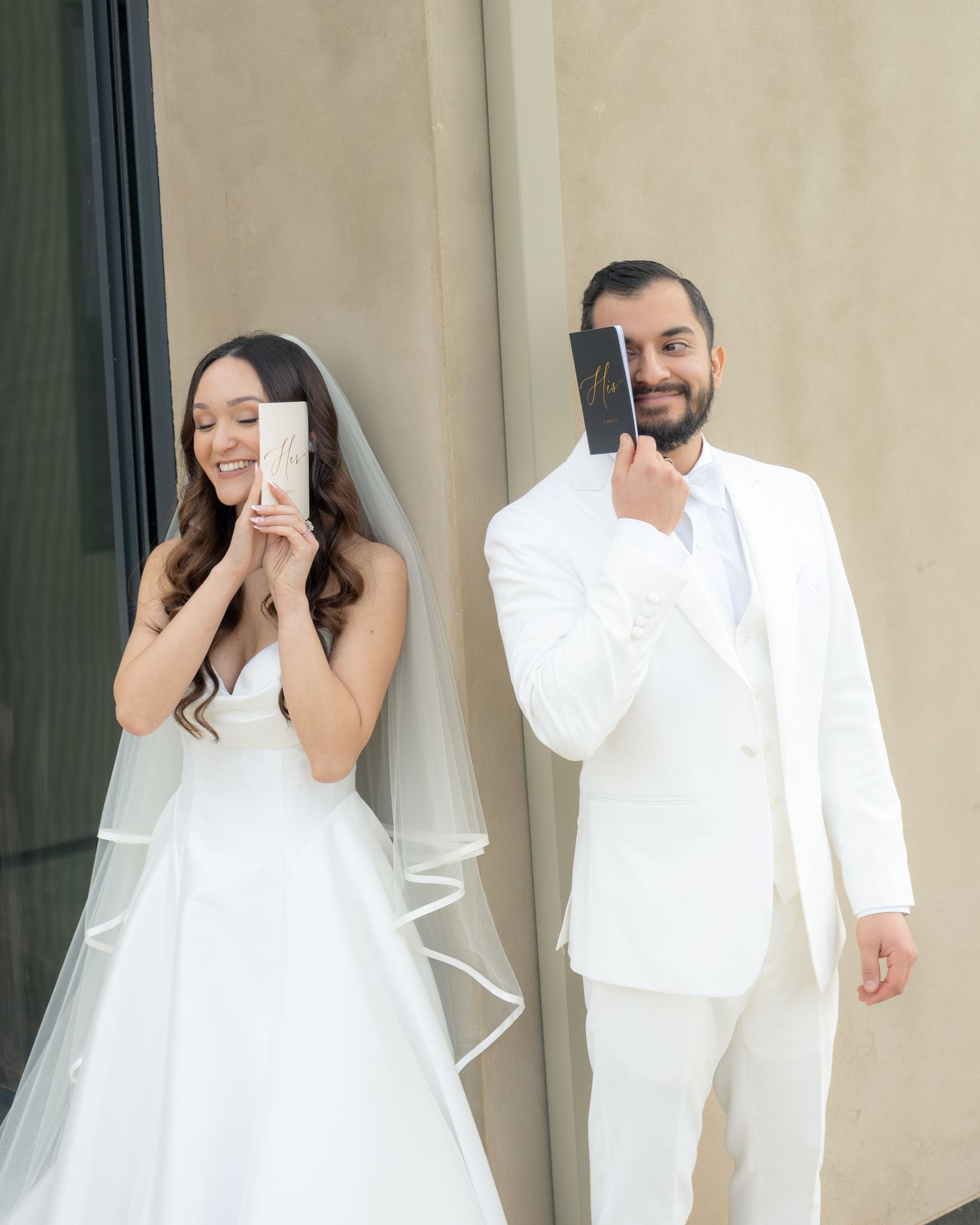 A bride and groom in white attire smile while holding books over their faces on their wedding day.