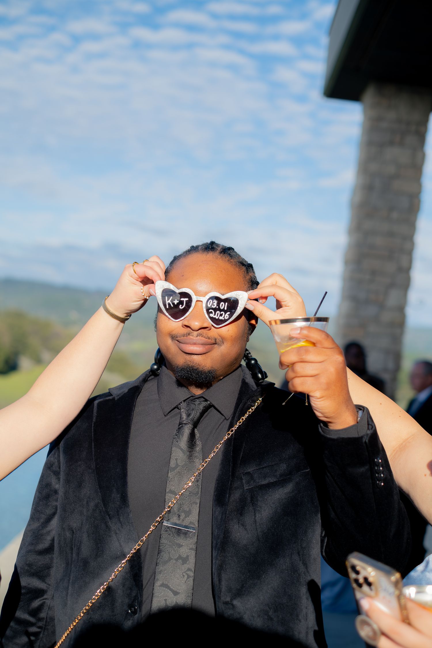 Wedding guest wearing heart-shaped novelty sunglasses with initials holds drink at reception.