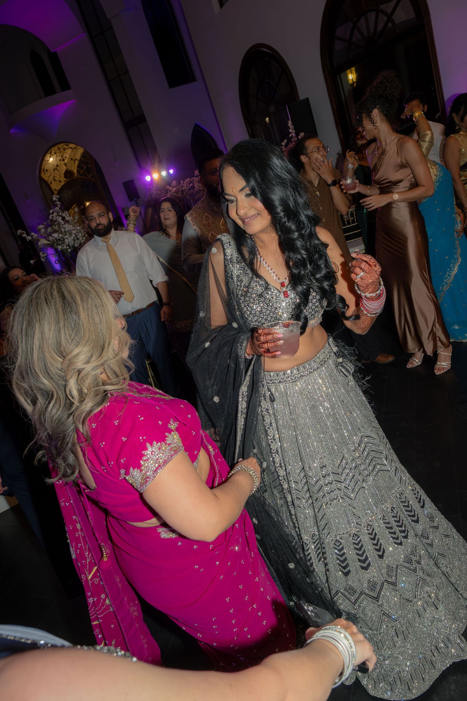 Two women in elegant Indian attire, one in grey lehenga and one in pink saree, share a moment on a festive dance floor.