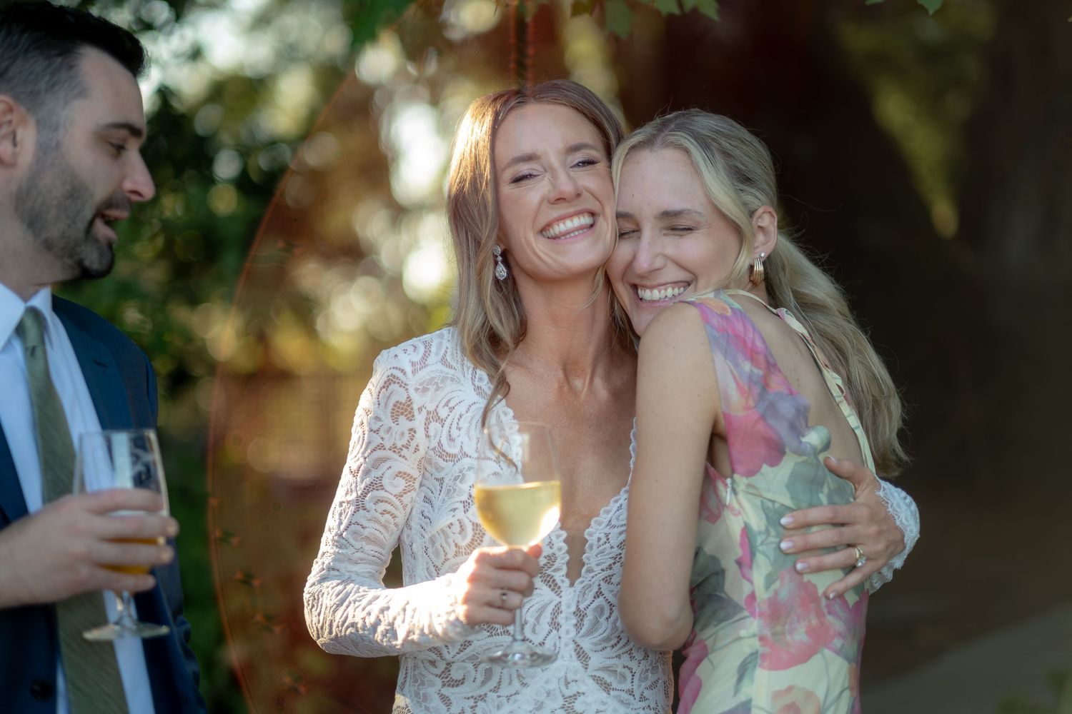 Bride in lace gown laughing and embracing a guest in a floral dress at a wedding reception.
