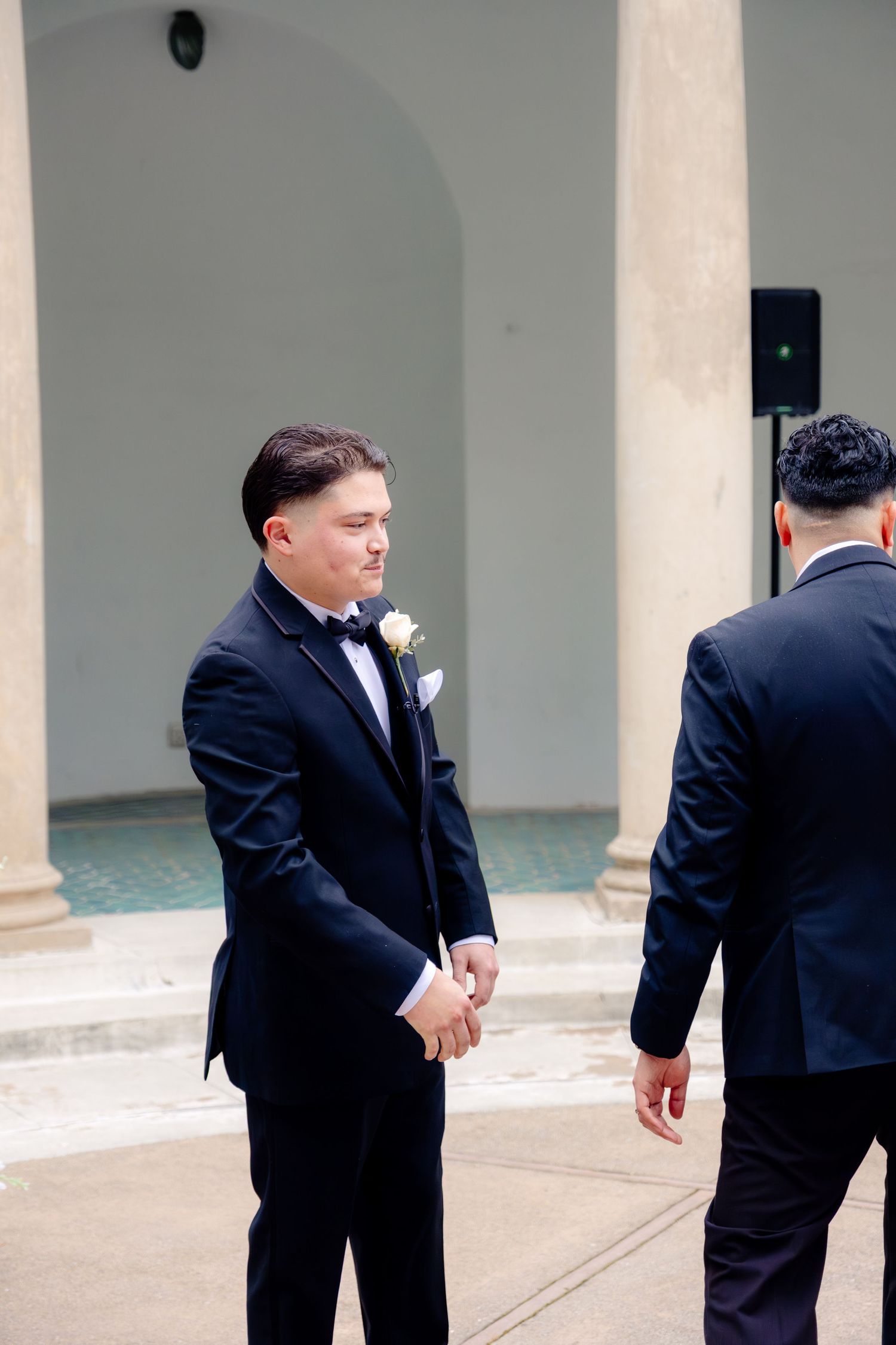 Groom in black tuxedo with white boutonniere stands outdoors at wedding venue with columns.