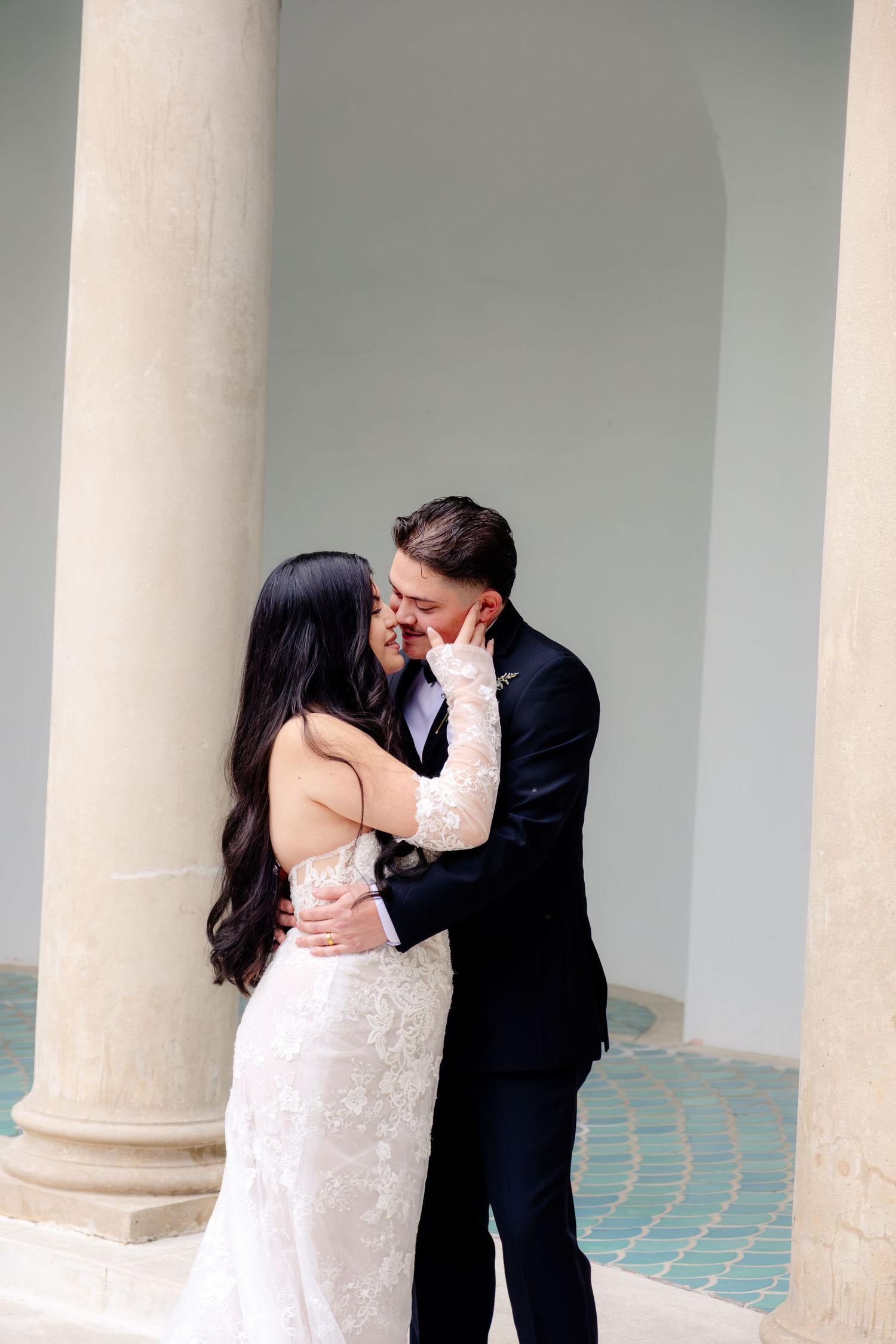 Newlyweds share a tender kiss between elegant white columns during their outdoor wedding photo session.