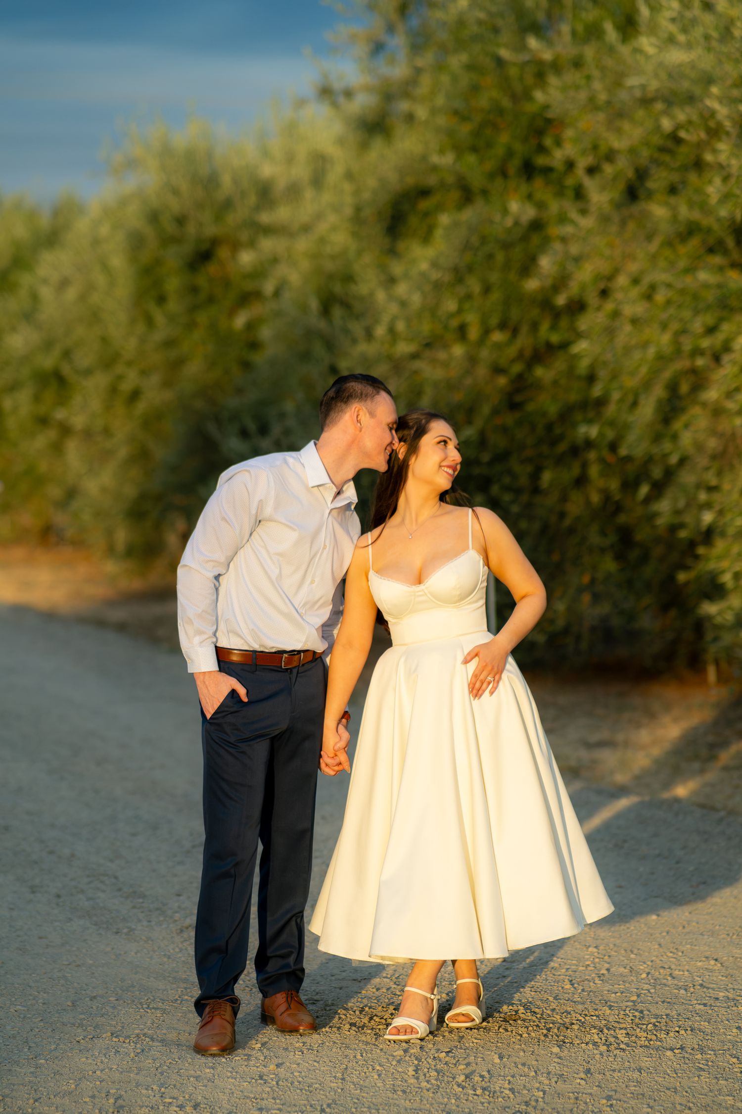 Couple holding hands on a gravel path lined with olive trees, groom kissing bride's temple in golden hour light.