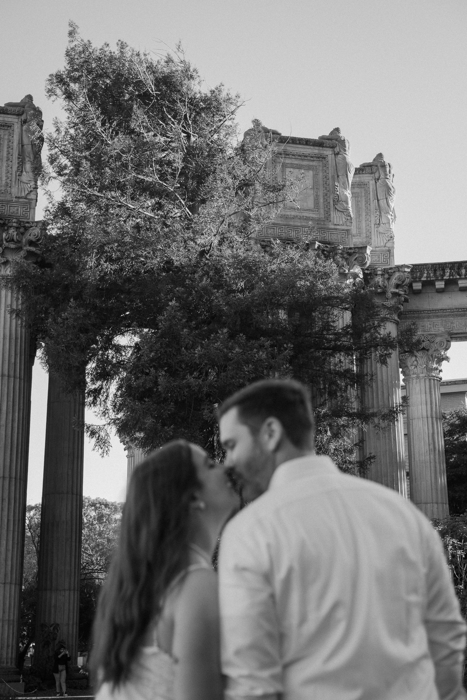 Black and white photo of a couple kissing in front of ancient Roman-style columns and a large tree.