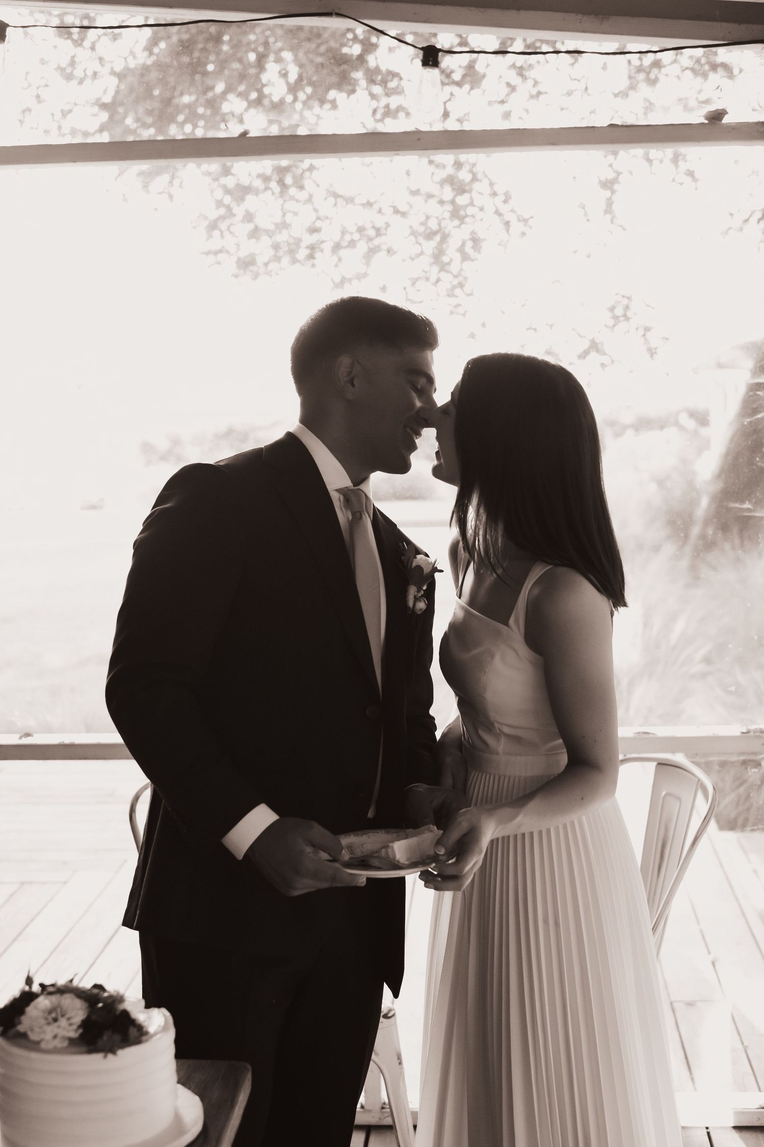 Black and white romantic photo of bride and groom nearly kissing beside their wedding cake.