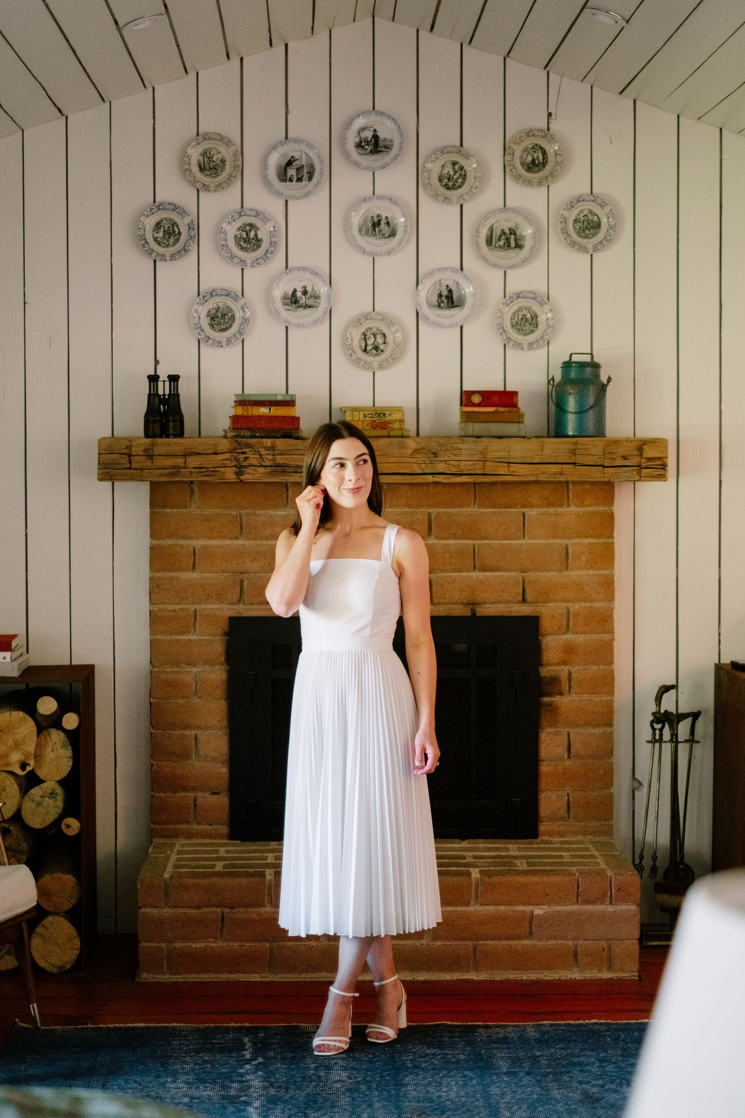 Bride in white pleated midi dress stands before brick fireplace with decorative plates on wall.