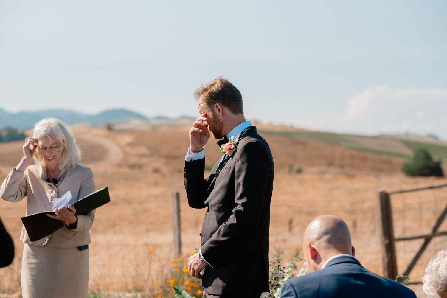 Emotional groom wiping tears during outdoor wedding ceremony in scenic countryside setting.