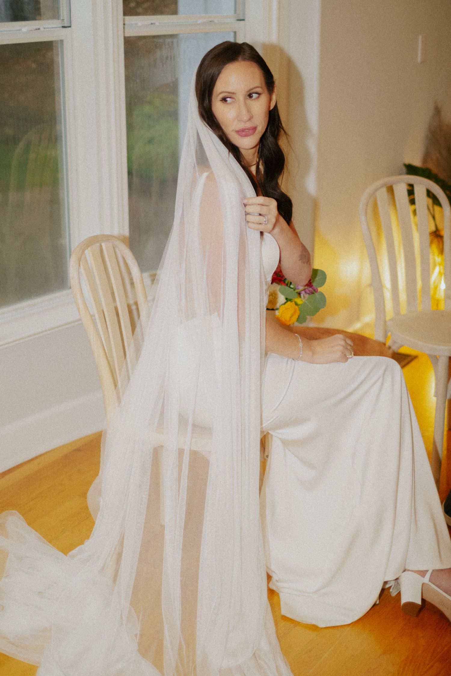 Bride in white gown and flowing veil sits gracefully by a sunlit window holding her bouquet.