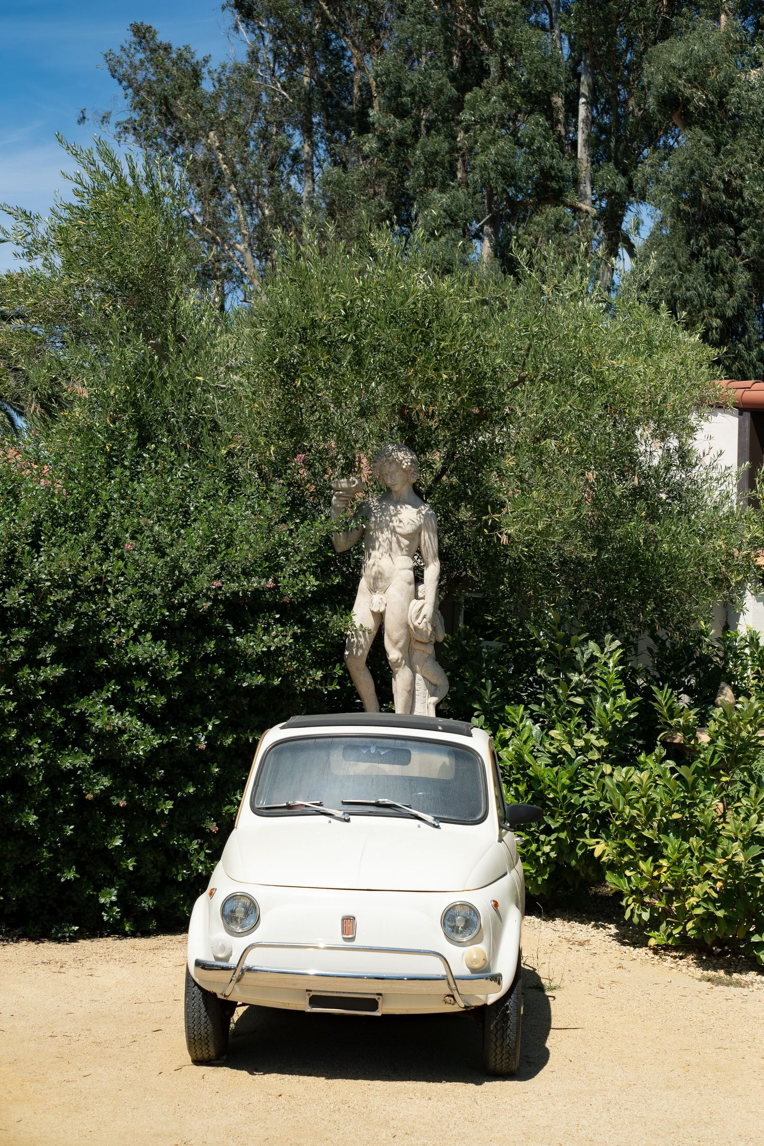 Vintage white Fiat 500 parked in front of a classical stone statue surrounded by lush greenery.