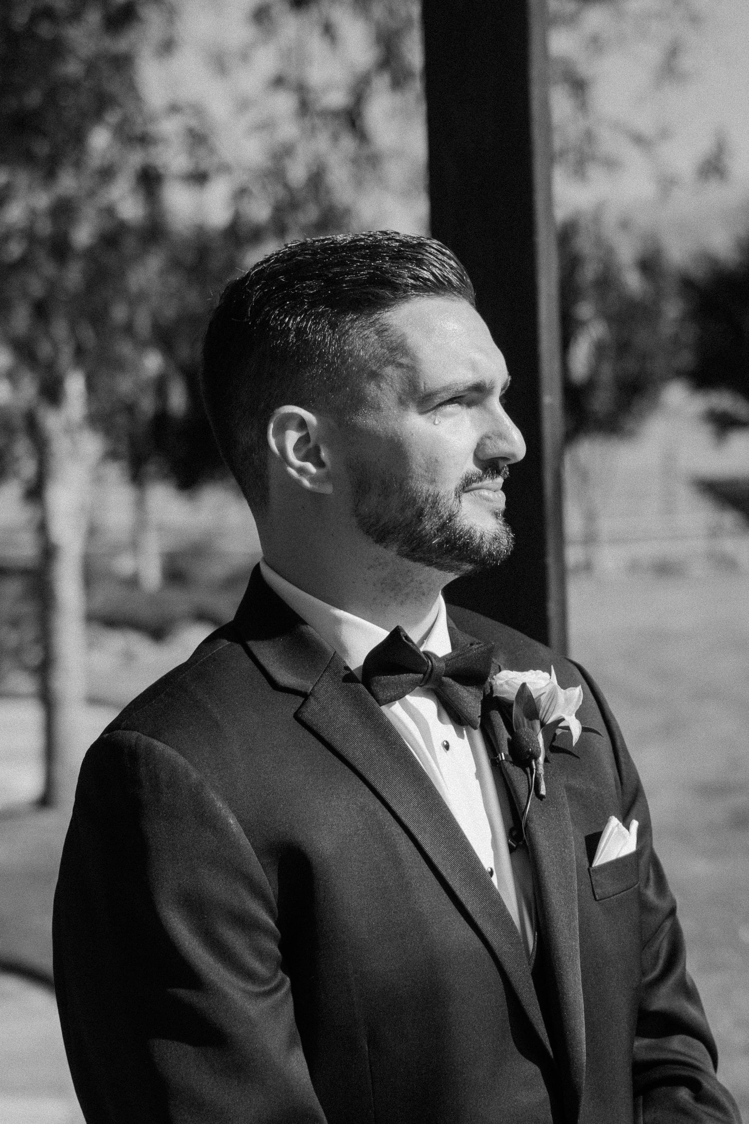 Black and white portrait of a groom in a tuxedo with bow tie and boutonniere at an outdoor wedding.