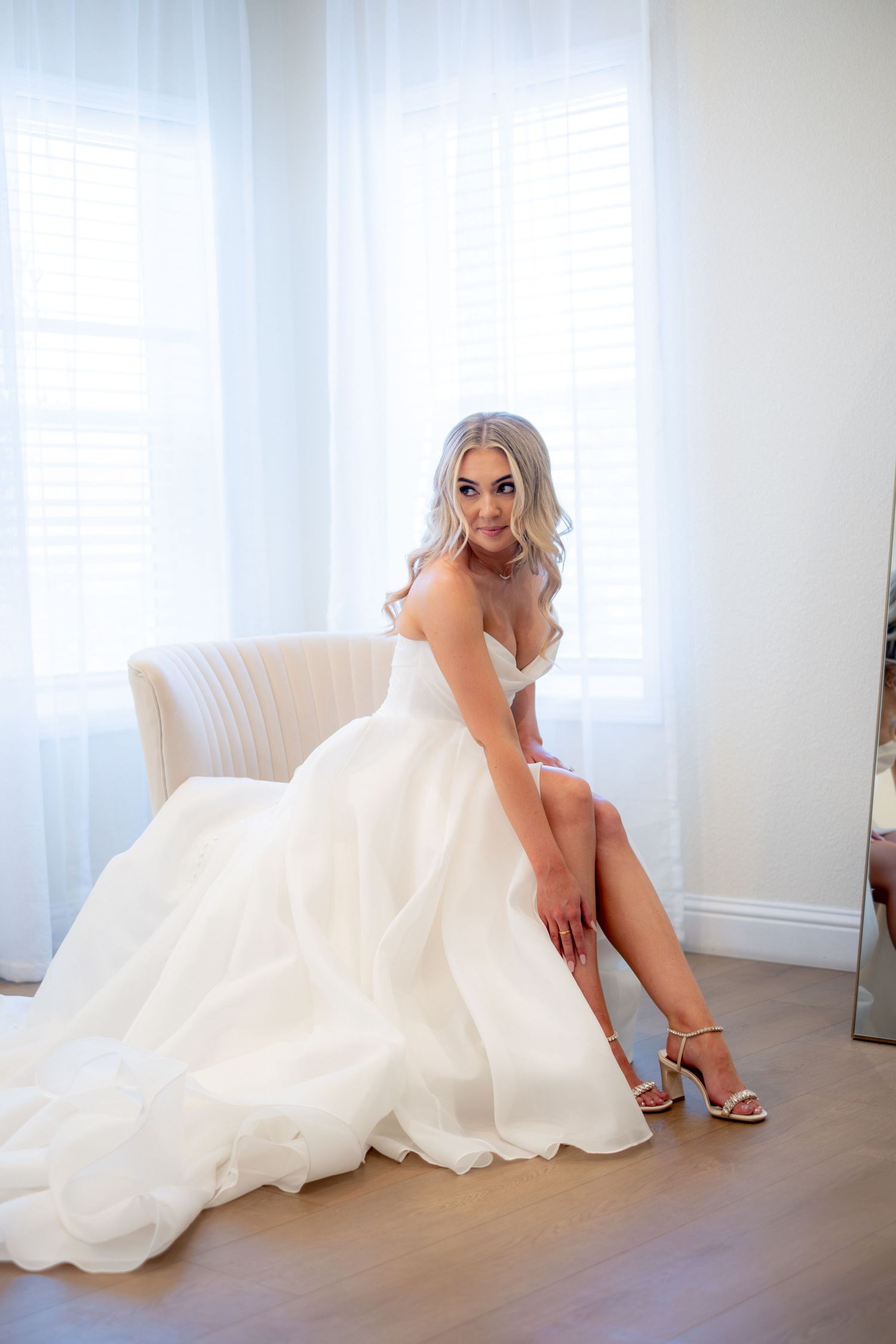 Elegant bride in a strapless white ball gown sits in a chair adjusting her gold strappy heels.
