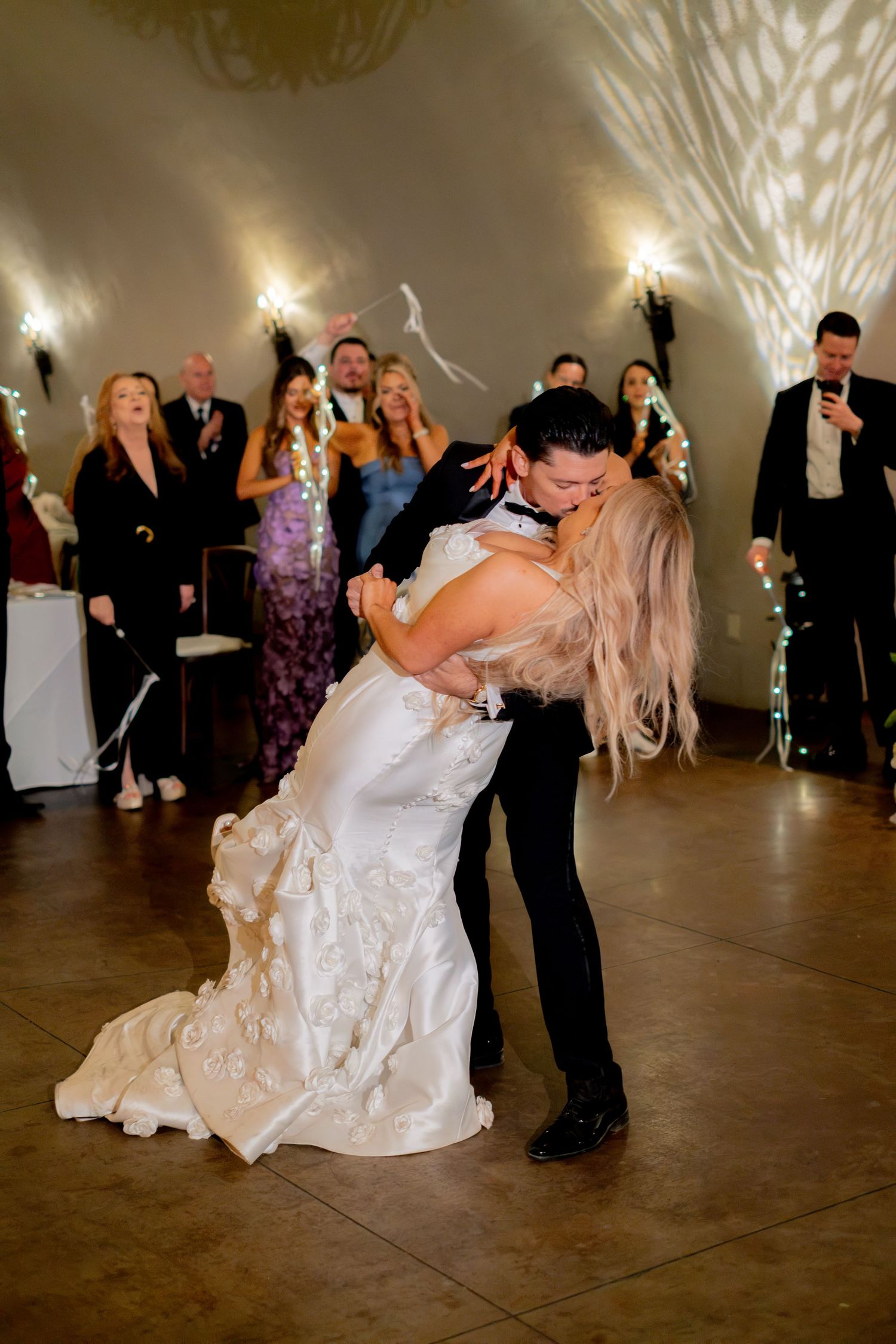Groom dips bride in white ruffled gown for romantic kiss on wedding reception dance floor.