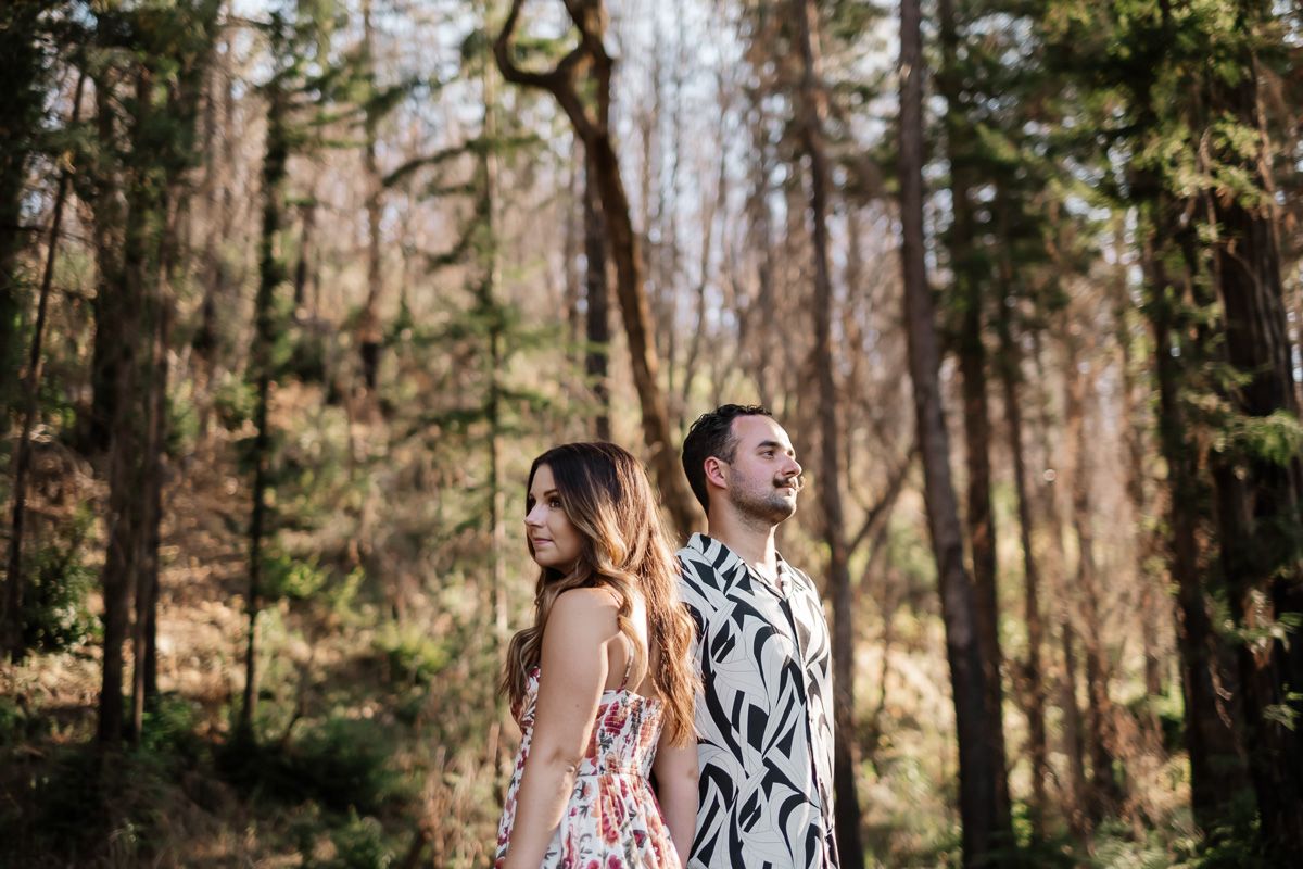Couple stands back to back smiling in a sunlit forest with tall trees behind them.