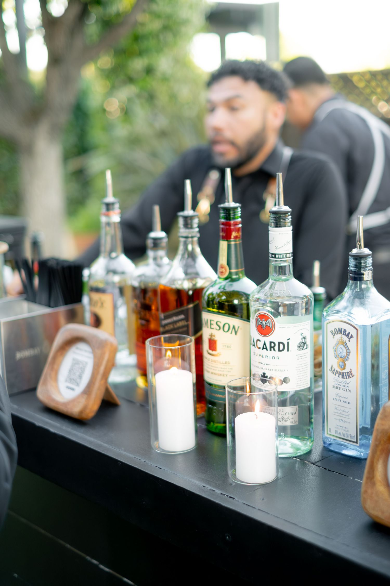 Wedding bar setup featuring Jameson, Bacardi, and Bombay Sapphire bottles with white candles and a bartender in background.
