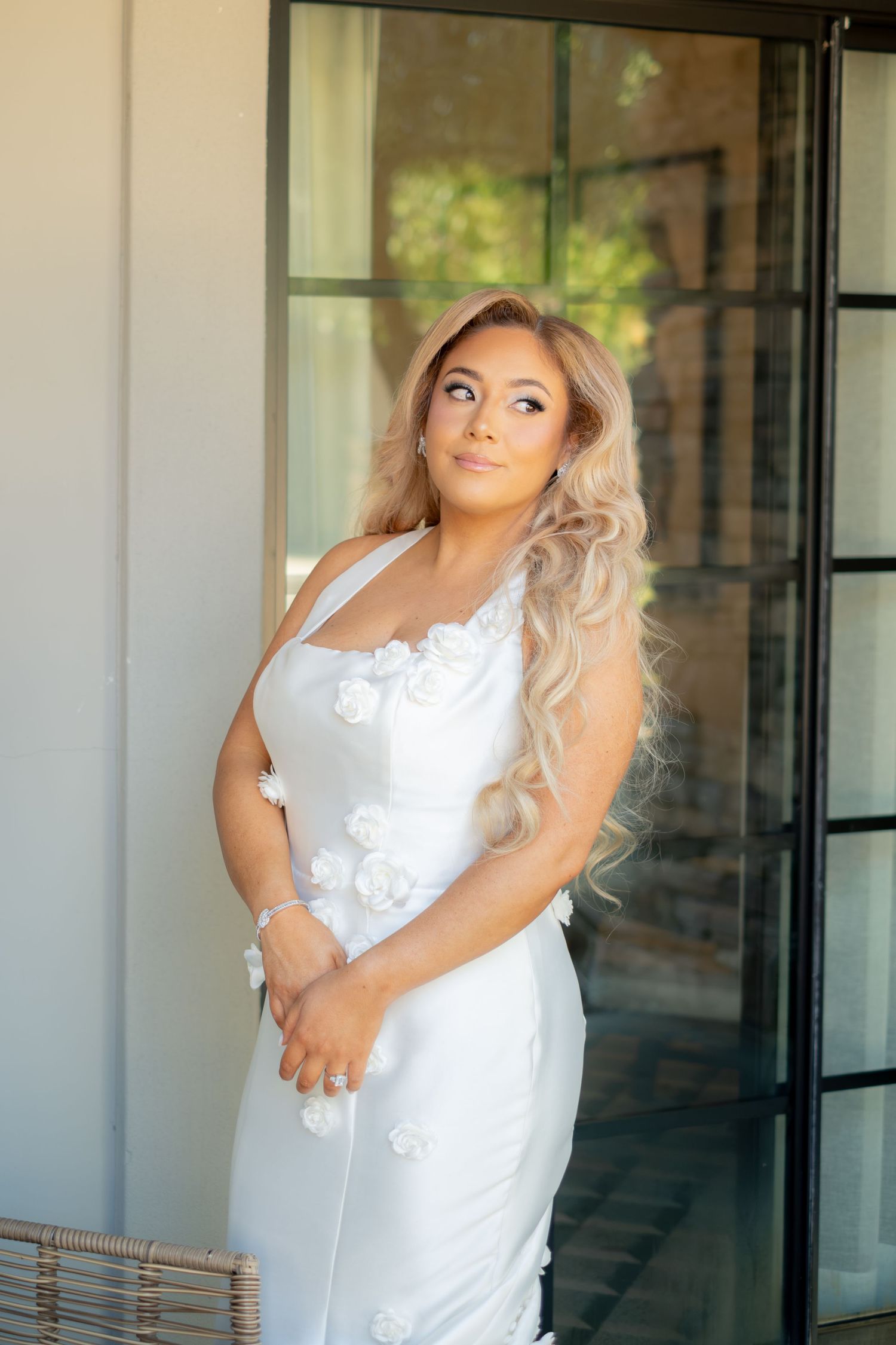 Elegant bride in a white floral applique gown with long wavy blonde hair posing by glass doors.