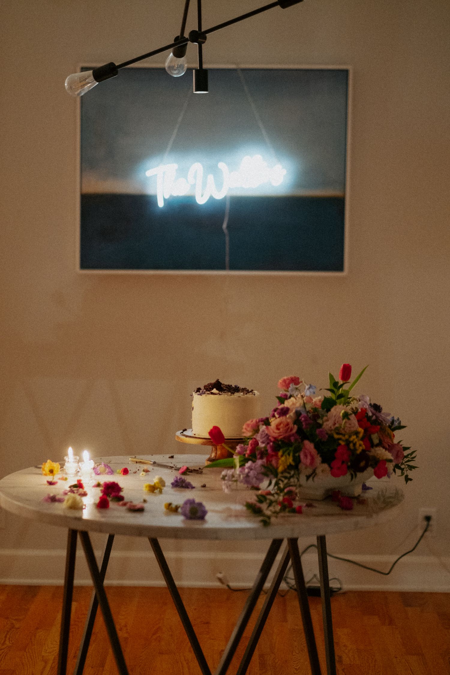 Wedding cake table with neon sign, candles, and vibrant floral arrangement against a painted wall.