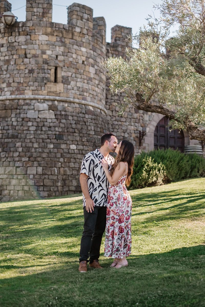 Couple sharing a romantic kiss on green lawn in front of a stone castle wall with arched entrance.