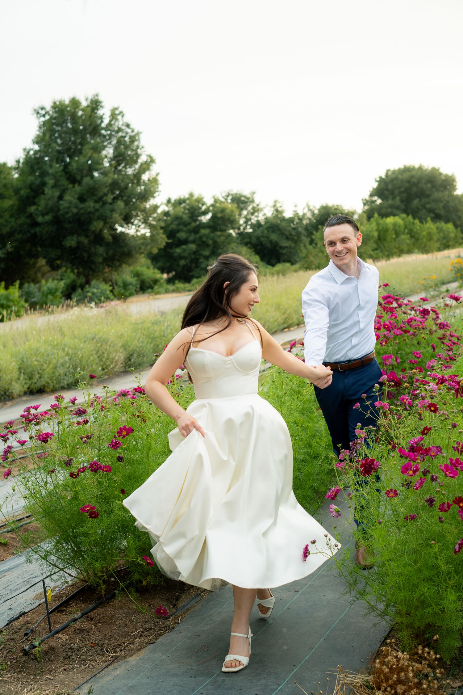 Bride in white tea-length dress twirling while holding hands with groom among pink cosmos flowers in a garden.