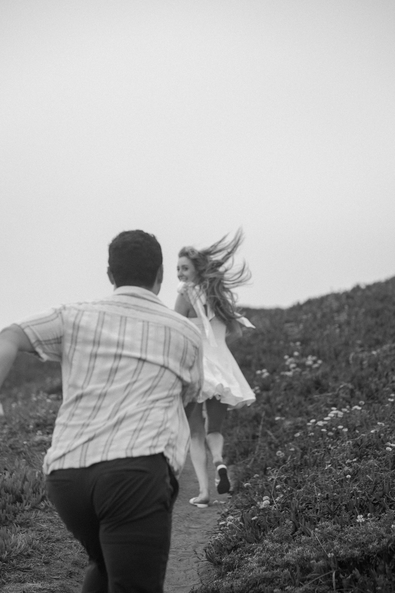 Black and white photo of woman in white dress running toward man on a hillside path.