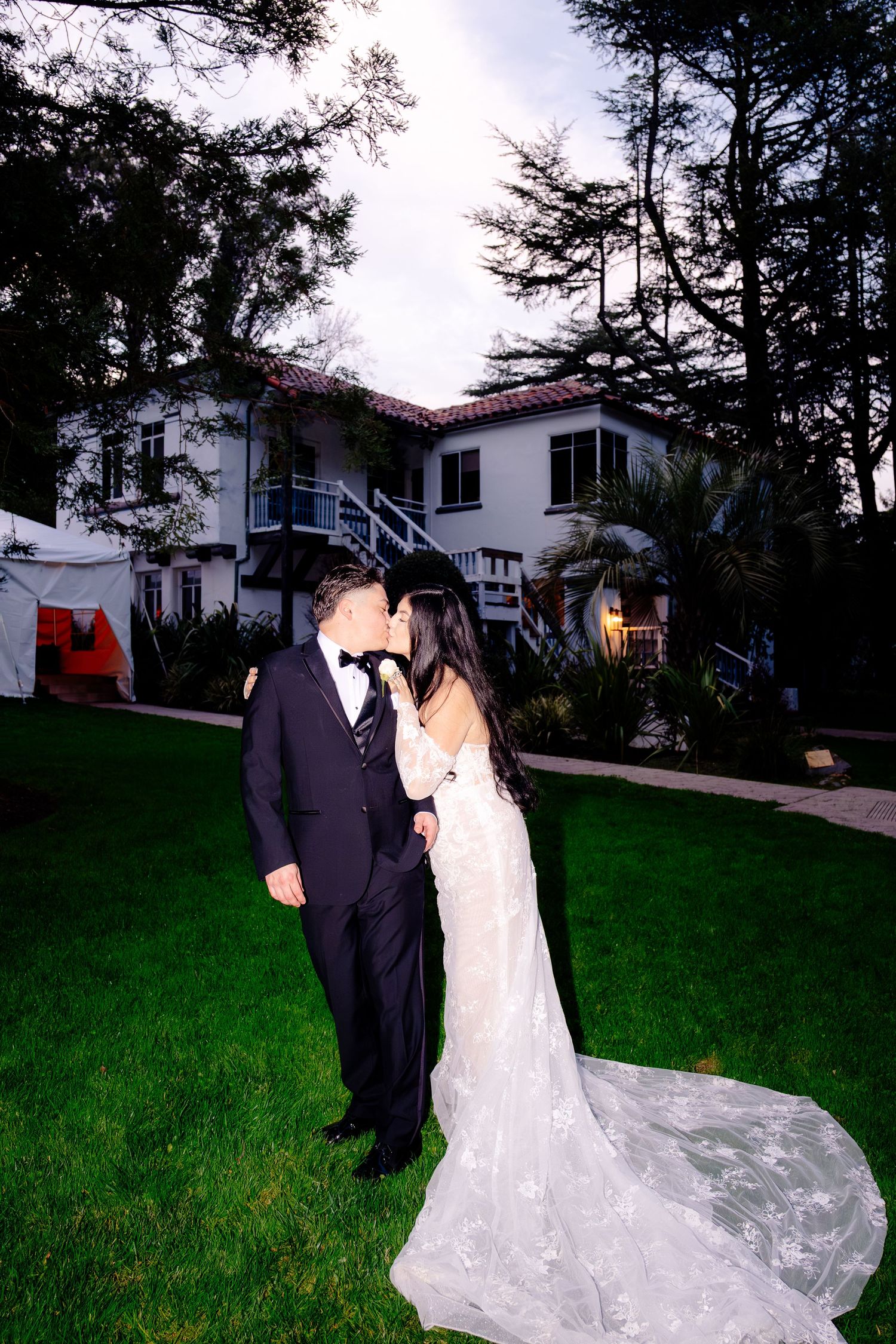 Bride and groom sharing a kiss on lush green lawn at dusk outside elegant white wedding venue.