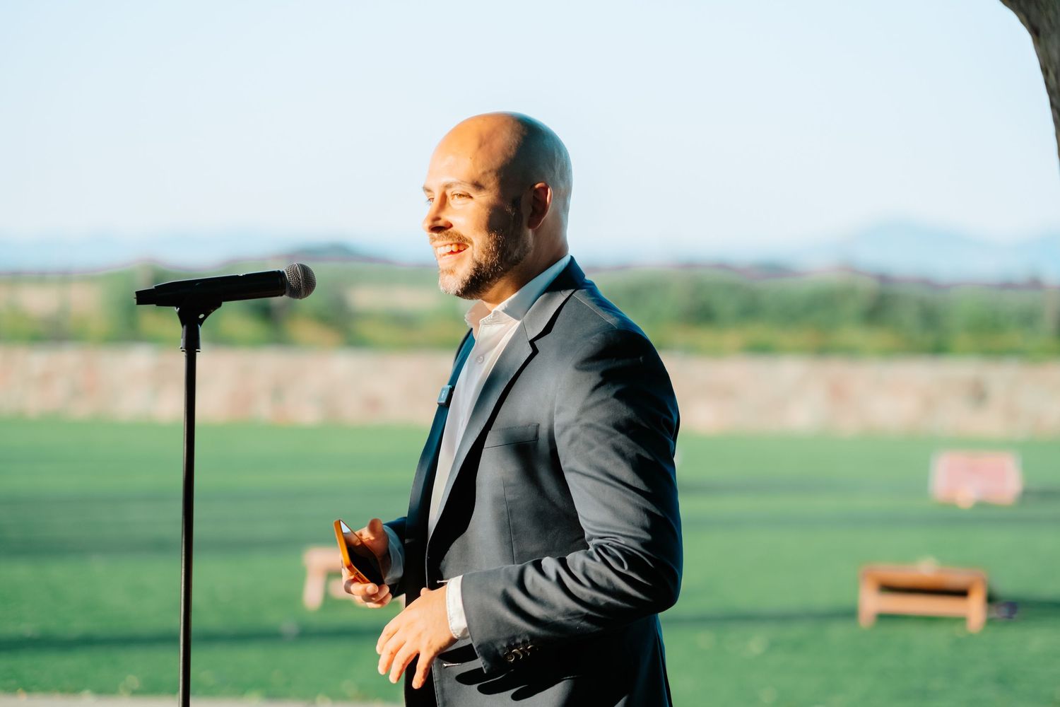 Smiling man in gray suit speaking at microphone during outdoor wedding reception with green field backdrop.