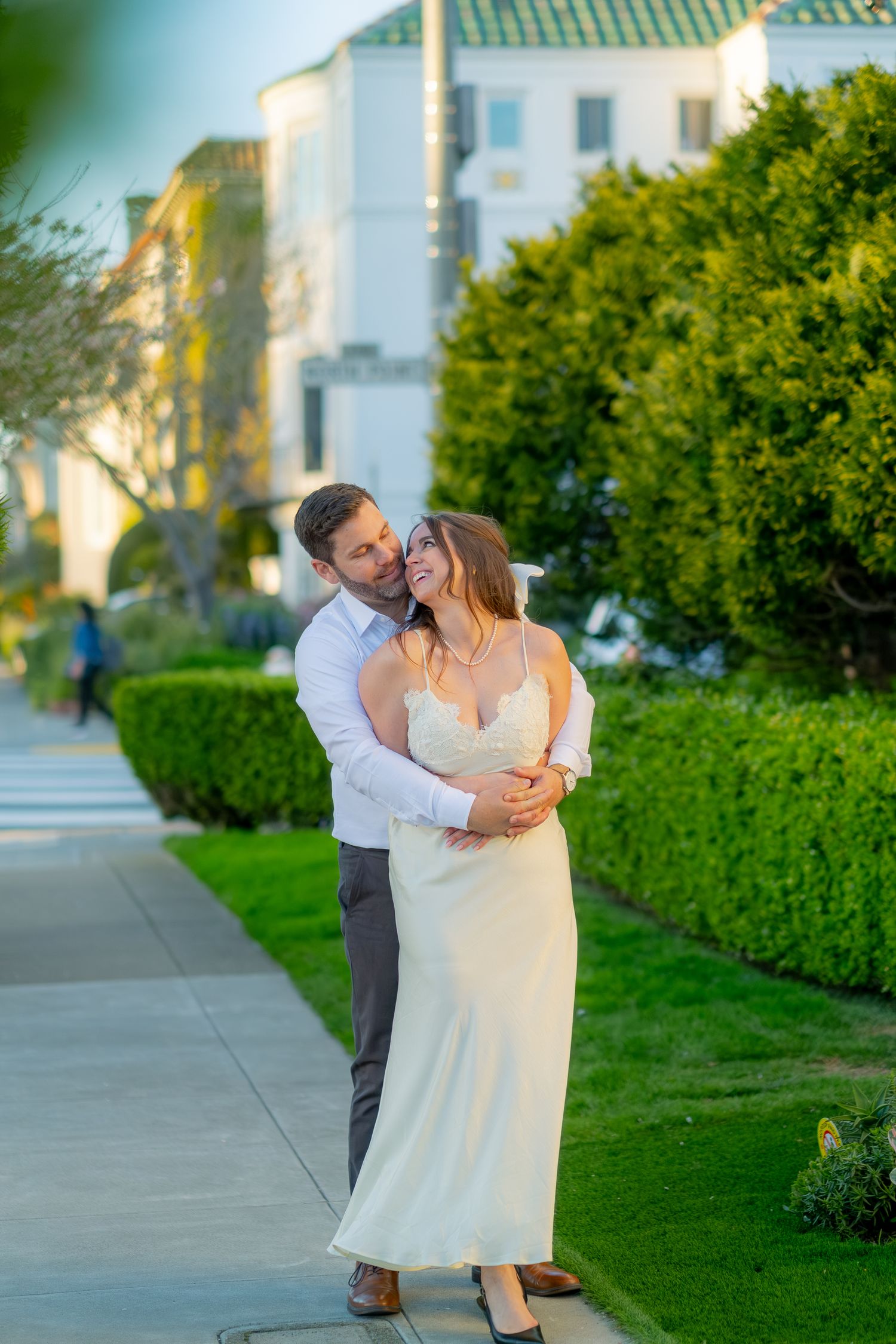 Couple embracing on a sunny sidewalk surrounded by lush green hedges in San Francisco.