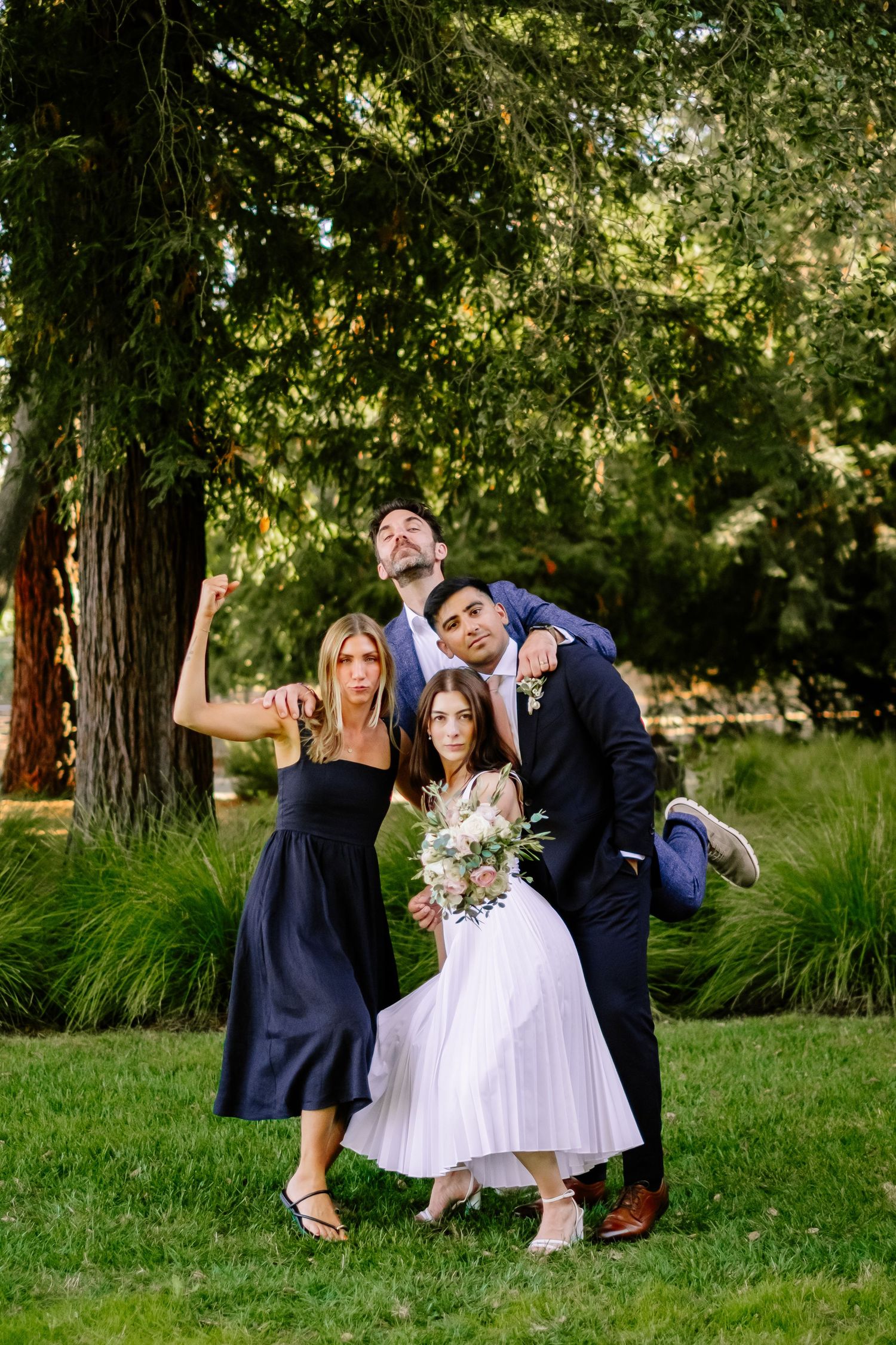 Wedding party poses playfully on green lawn under large tree with bride holding bouquet.