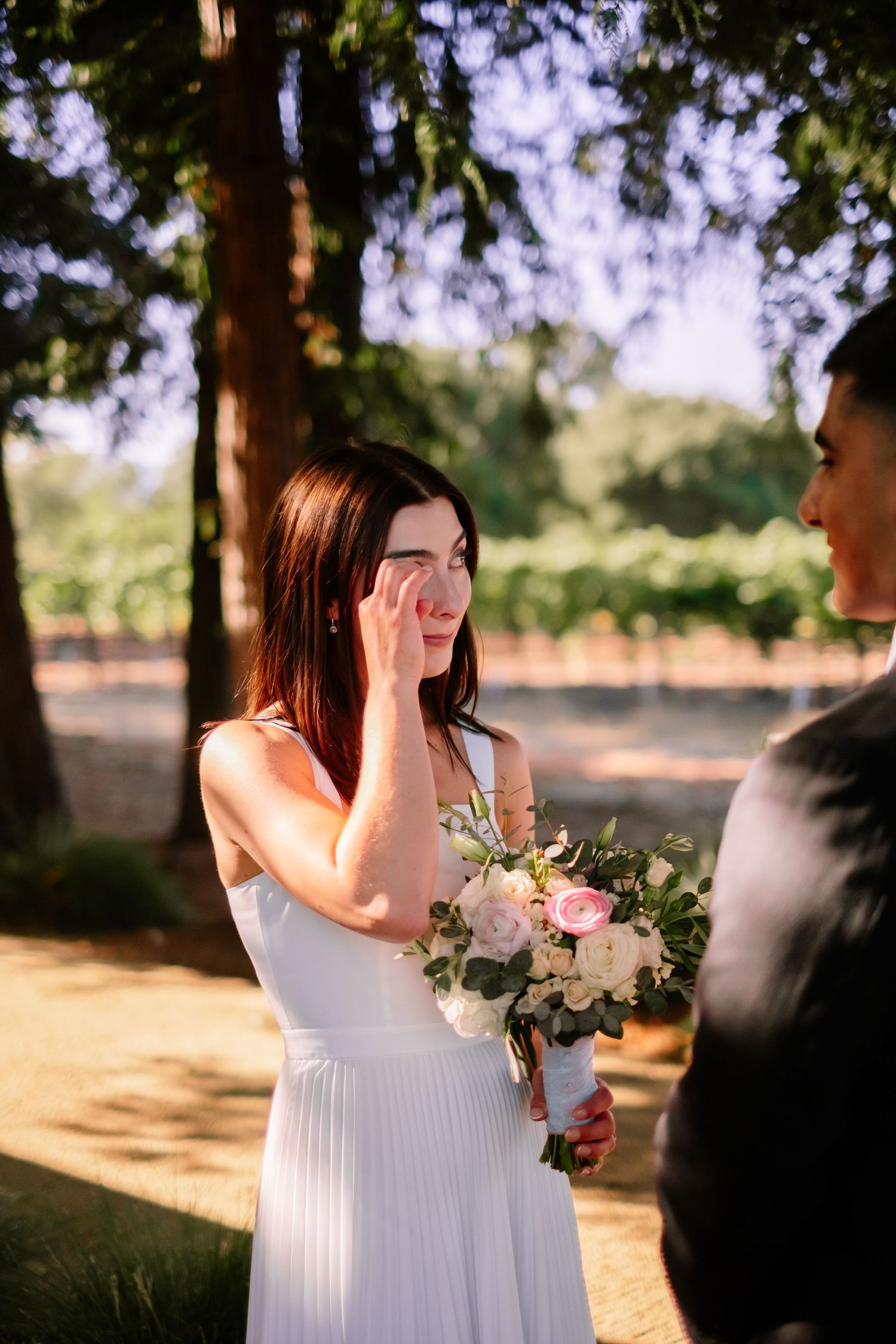 Tearful bride in white dress holding pink and white bouquet during outdoor wedding ceremony.