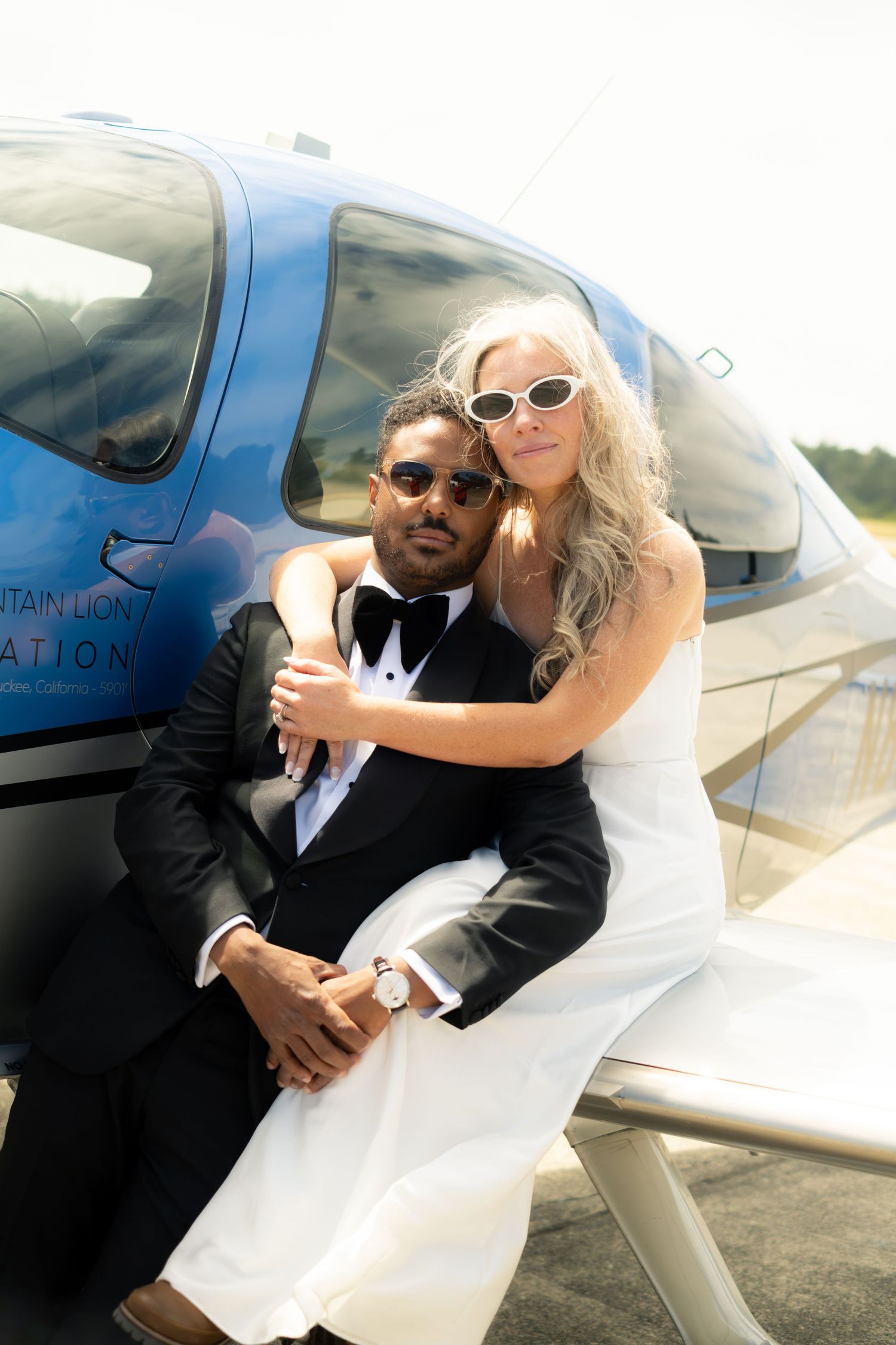 Elegant interracial couple in wedding attire posing by a blue helicopter on a sunny day.