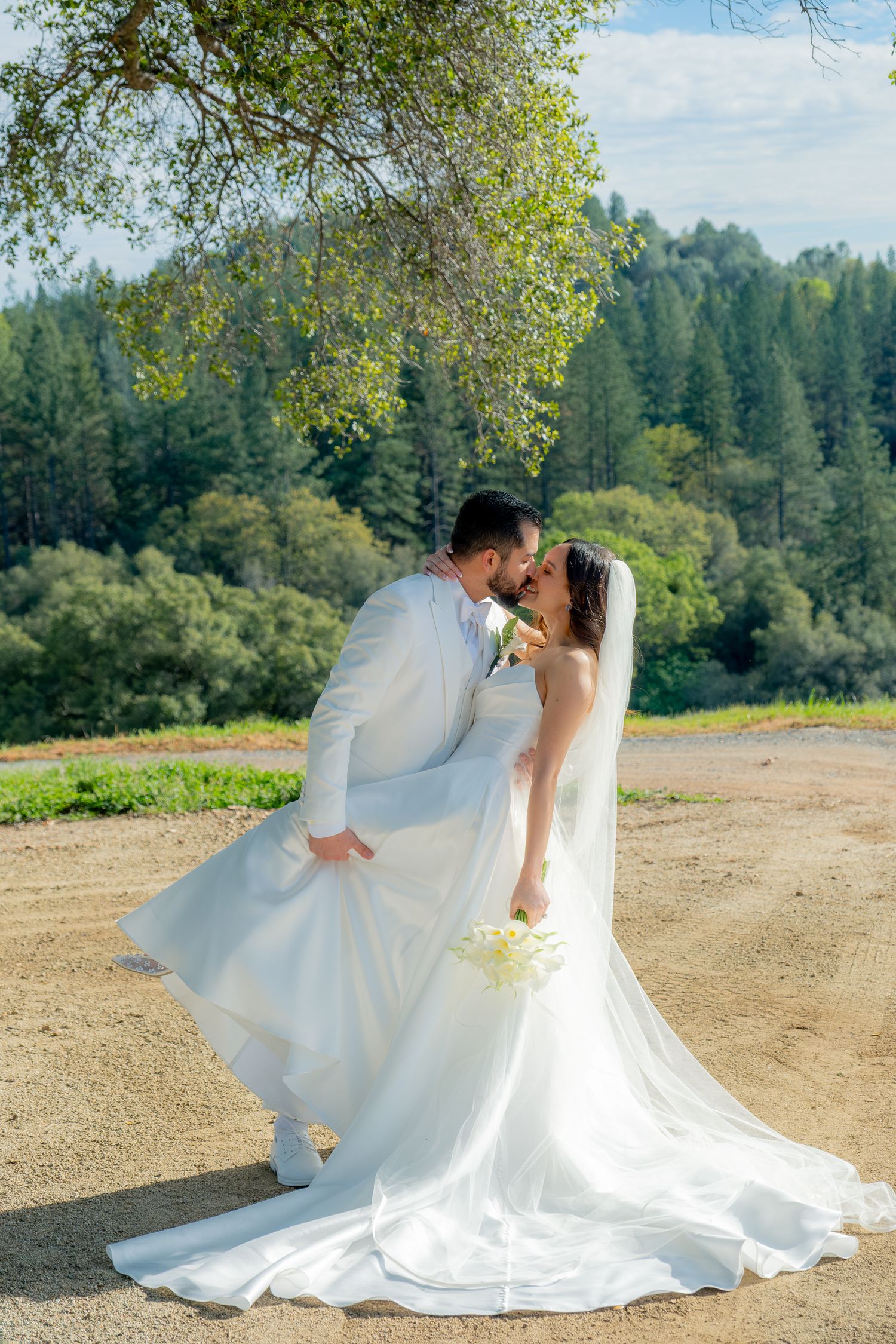 Bride and groom share a romantic kiss outdoors, bride in flowing white gown with veil, groom in white suit, forest backdrop.