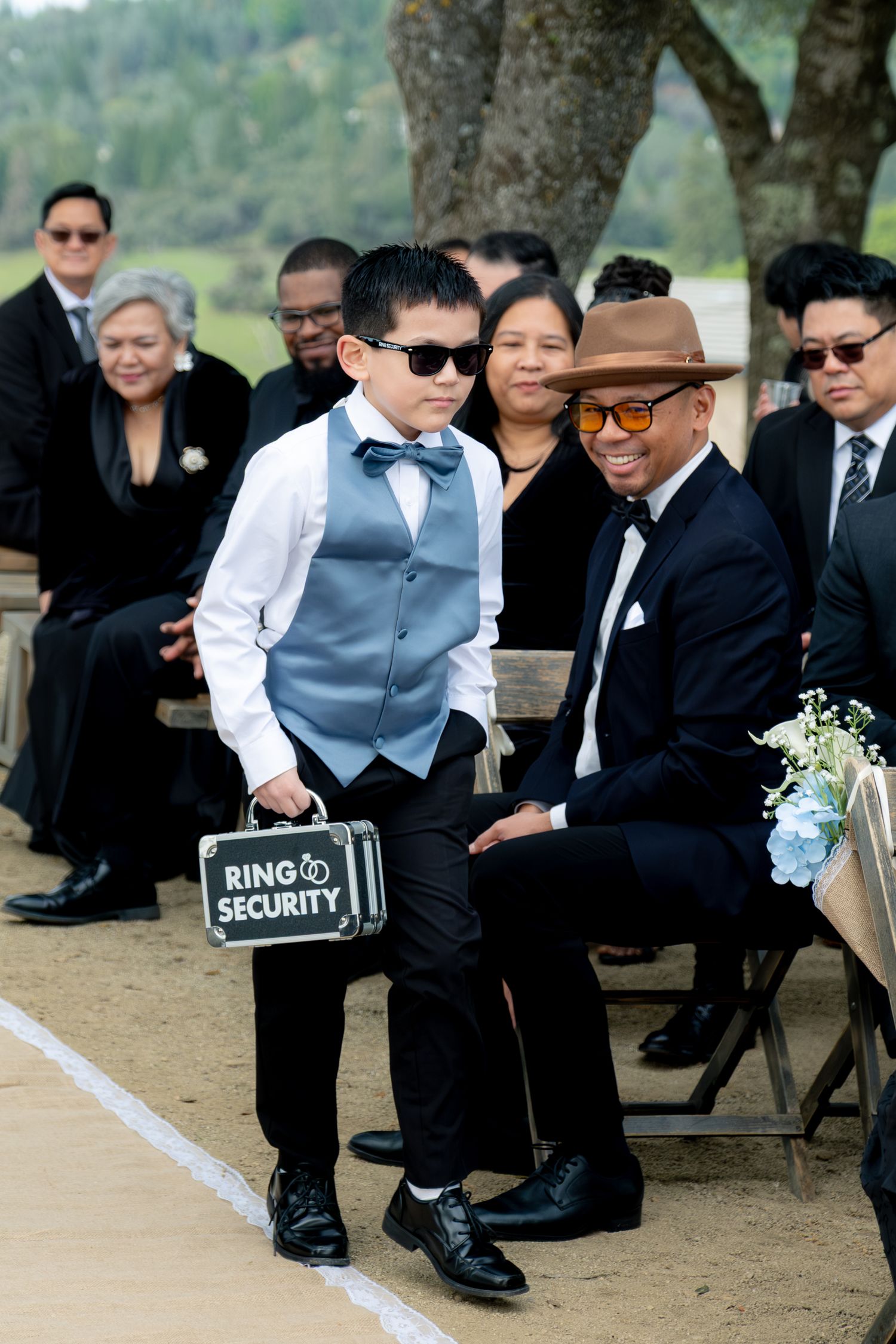 Adorable ring bearer in blue vest carries Ring Security briefcase down wedding aisle.