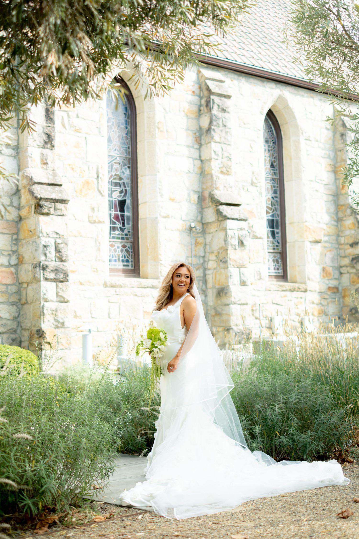 Radiant bride in white gown holding green bouquet outside stone church with stained glass.