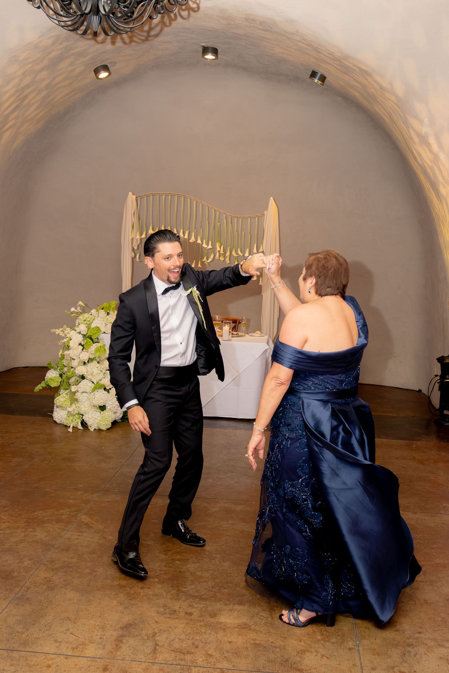 Groom dances joyfully with his mother in a navy gown under a stone arch at the reception.