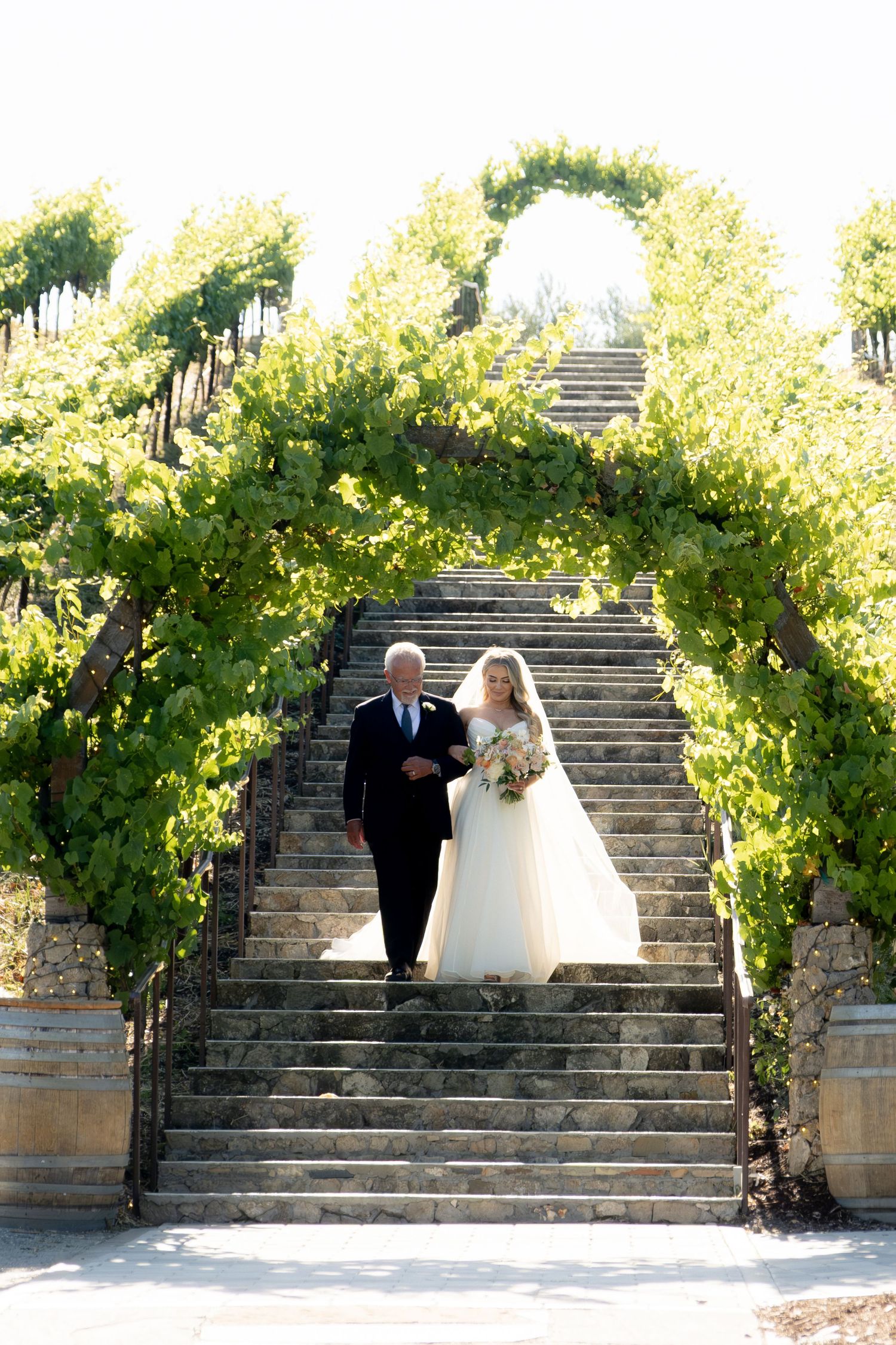Bride in white ball gown descends vine-covered stone staircase with her father on her wedding day.