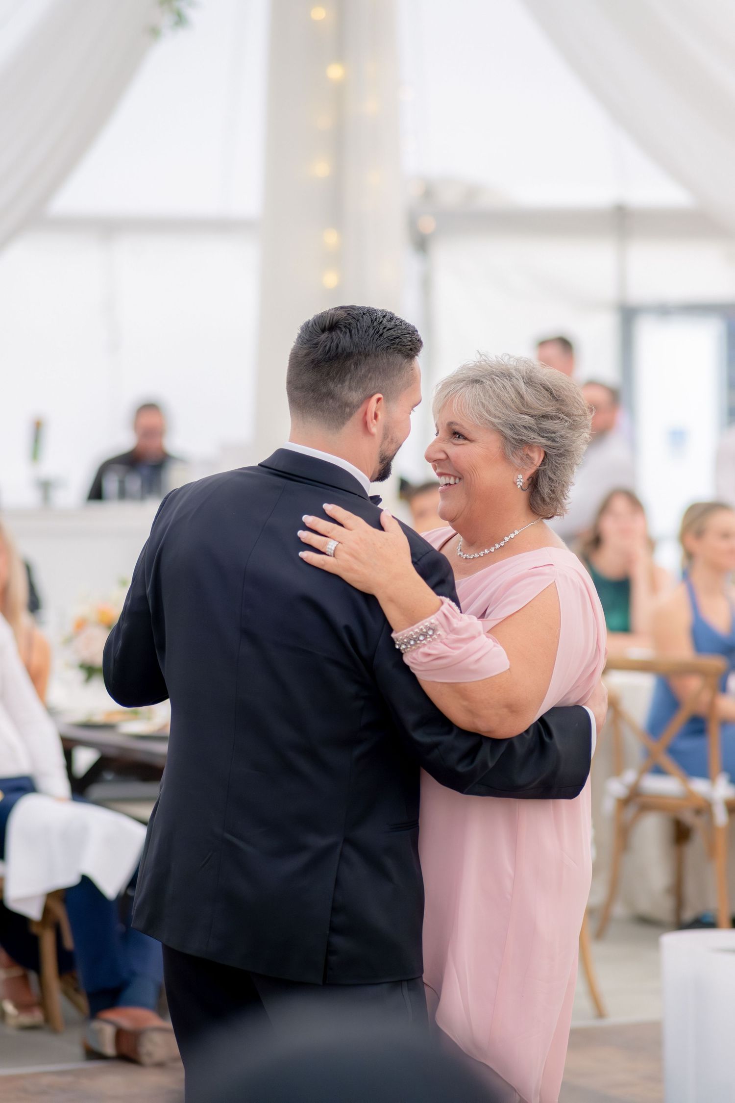 Groom sharing joyful mother-son dance under draped tent with string lights at wedding reception.