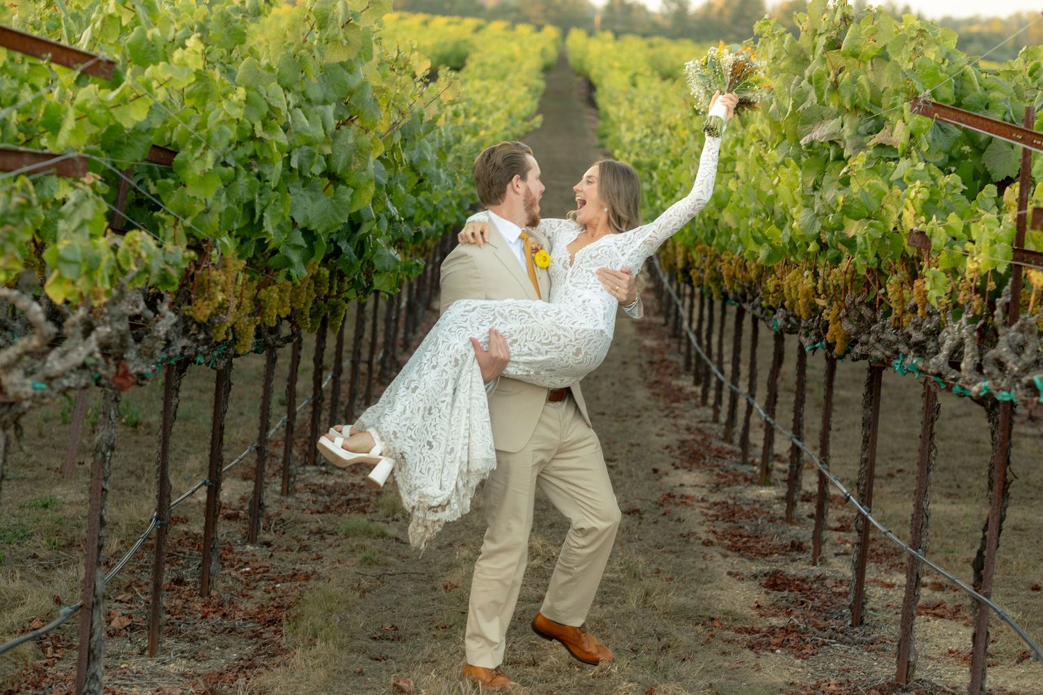 Groom carrying laughing bride through vineyard rows, both holding wine glasses at outdoor wedding.