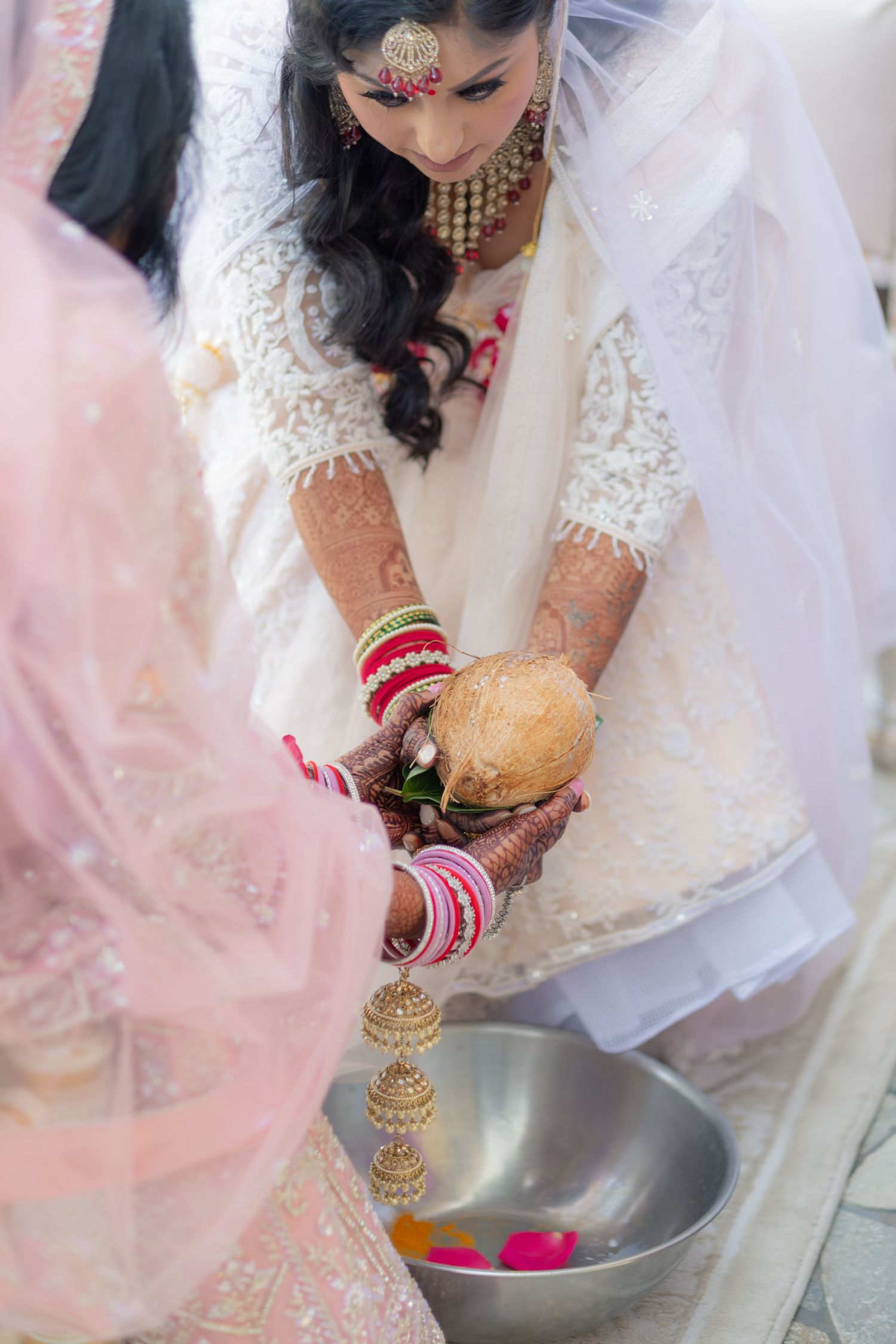 Indian bride with mehndi and red bangles performs traditional wedding ritual with coconut over silver bowl.