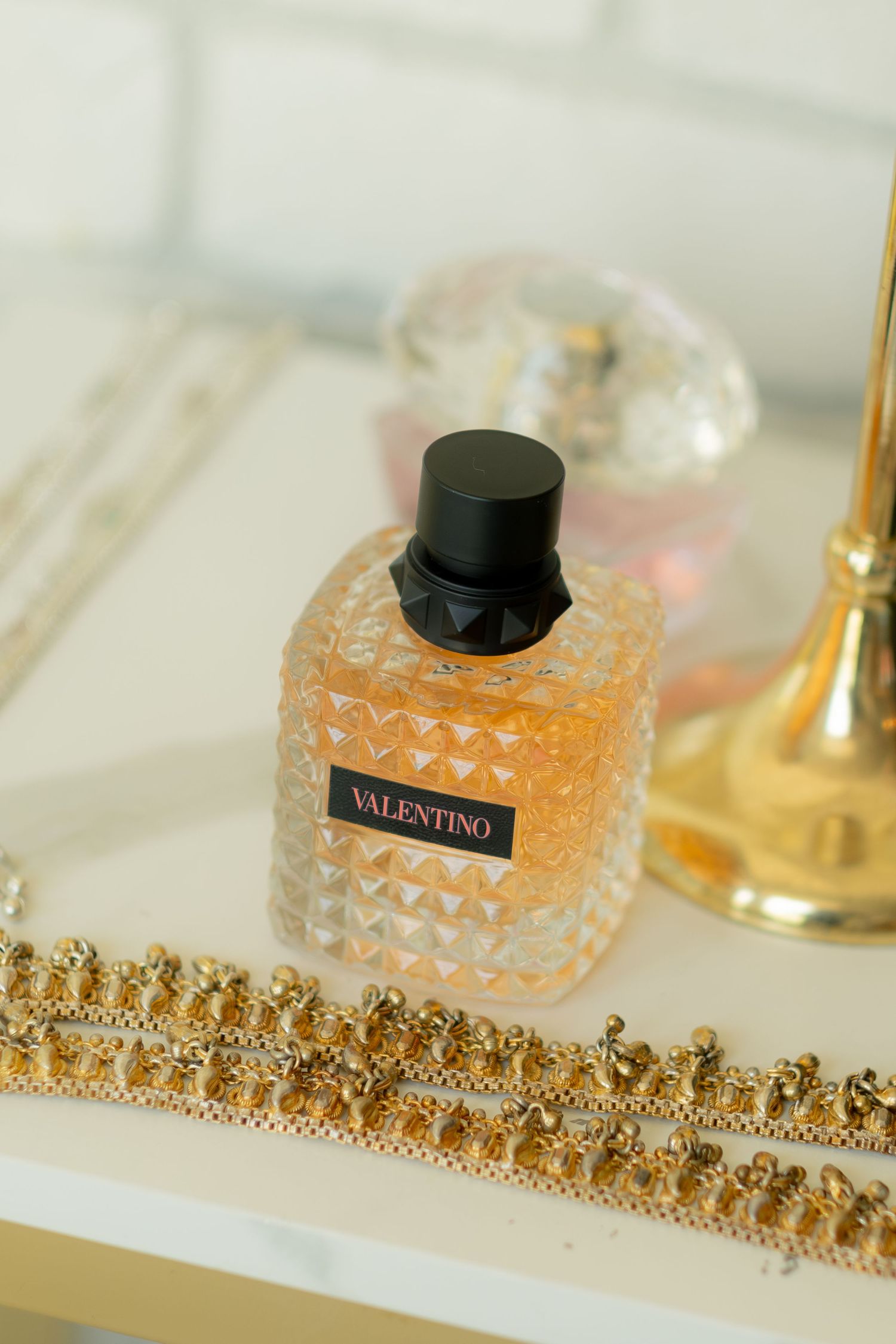 Valentino Born in Roma perfume bottle displayed with gold jewelry on white vanity surface.