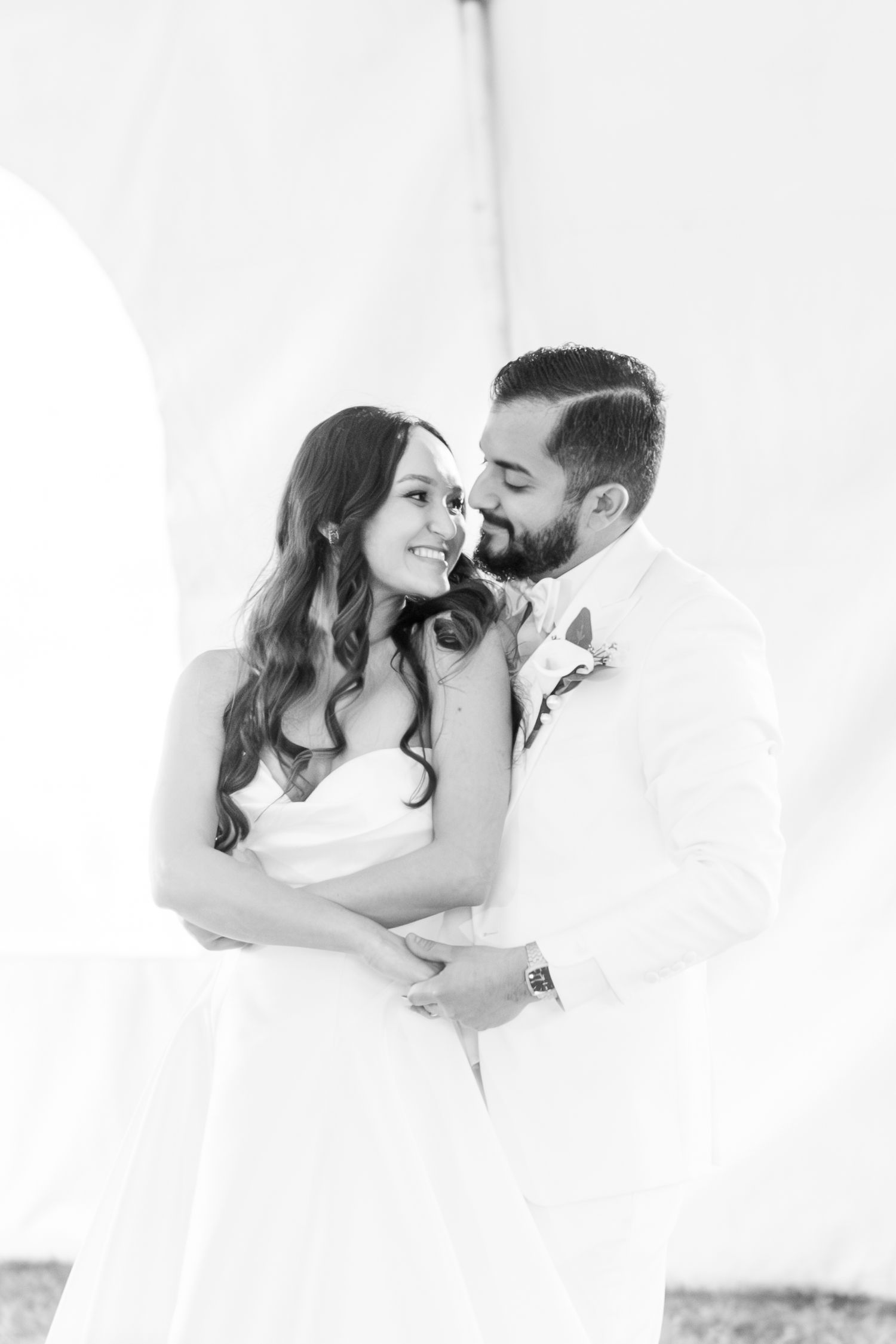 Black and white portrait of smiling bride and bearded groom embracing tenderly under white wedding tent.