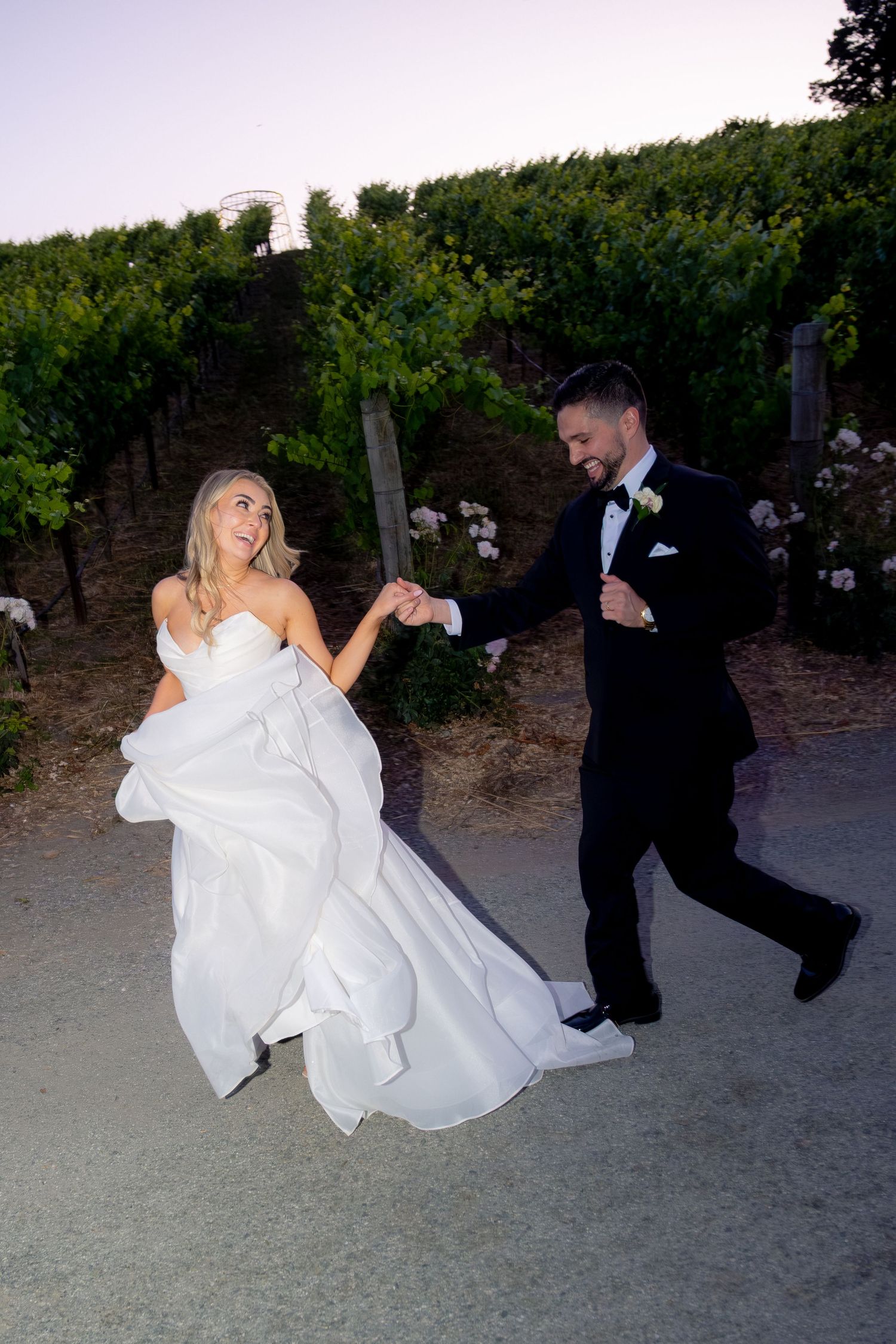 Bride and groom dance playfully on a path through a vineyard at dusk, holding hands.