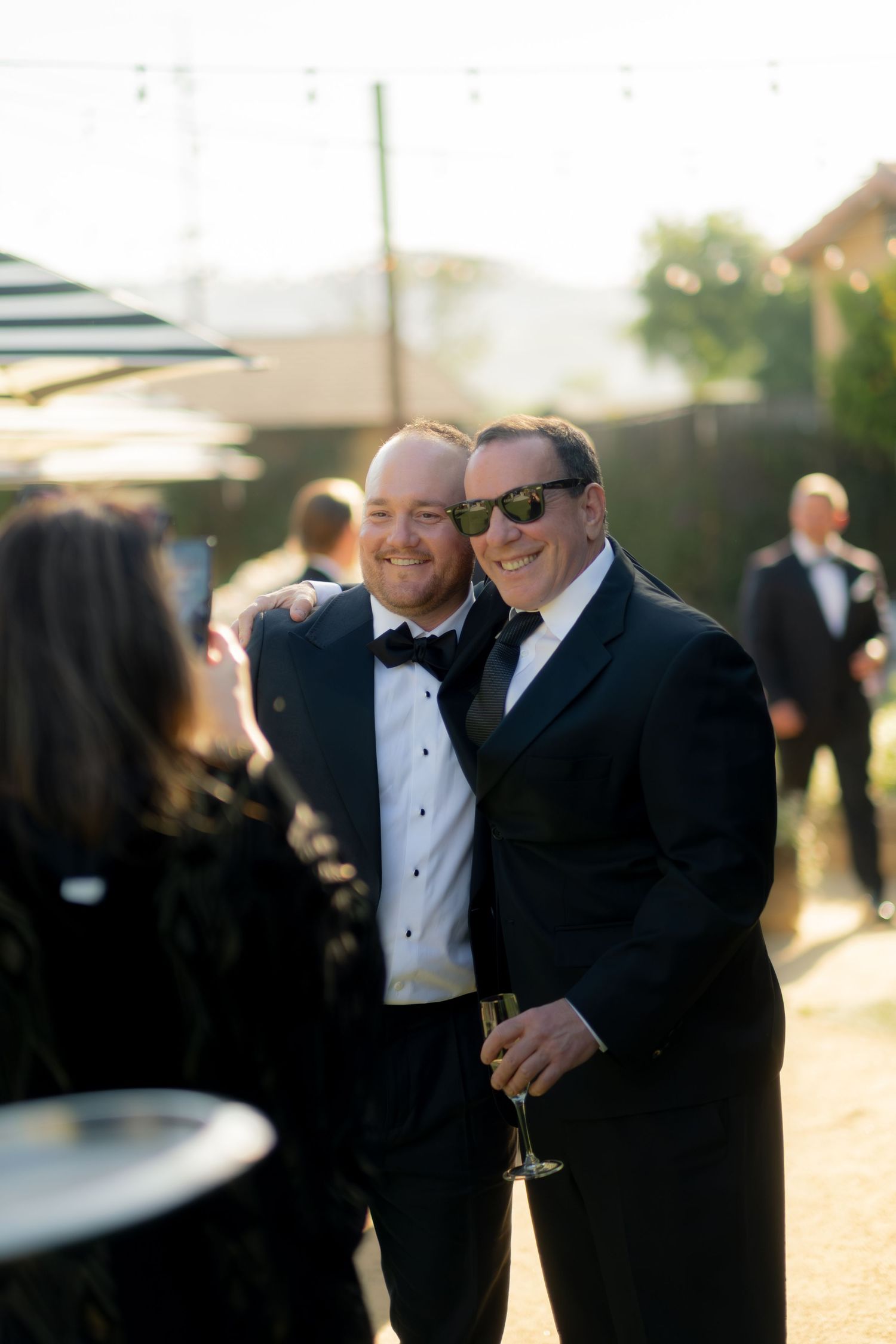 Two men in tuxedos smile for a photo at an outdoor wedding reception, one holding a champagne flute.