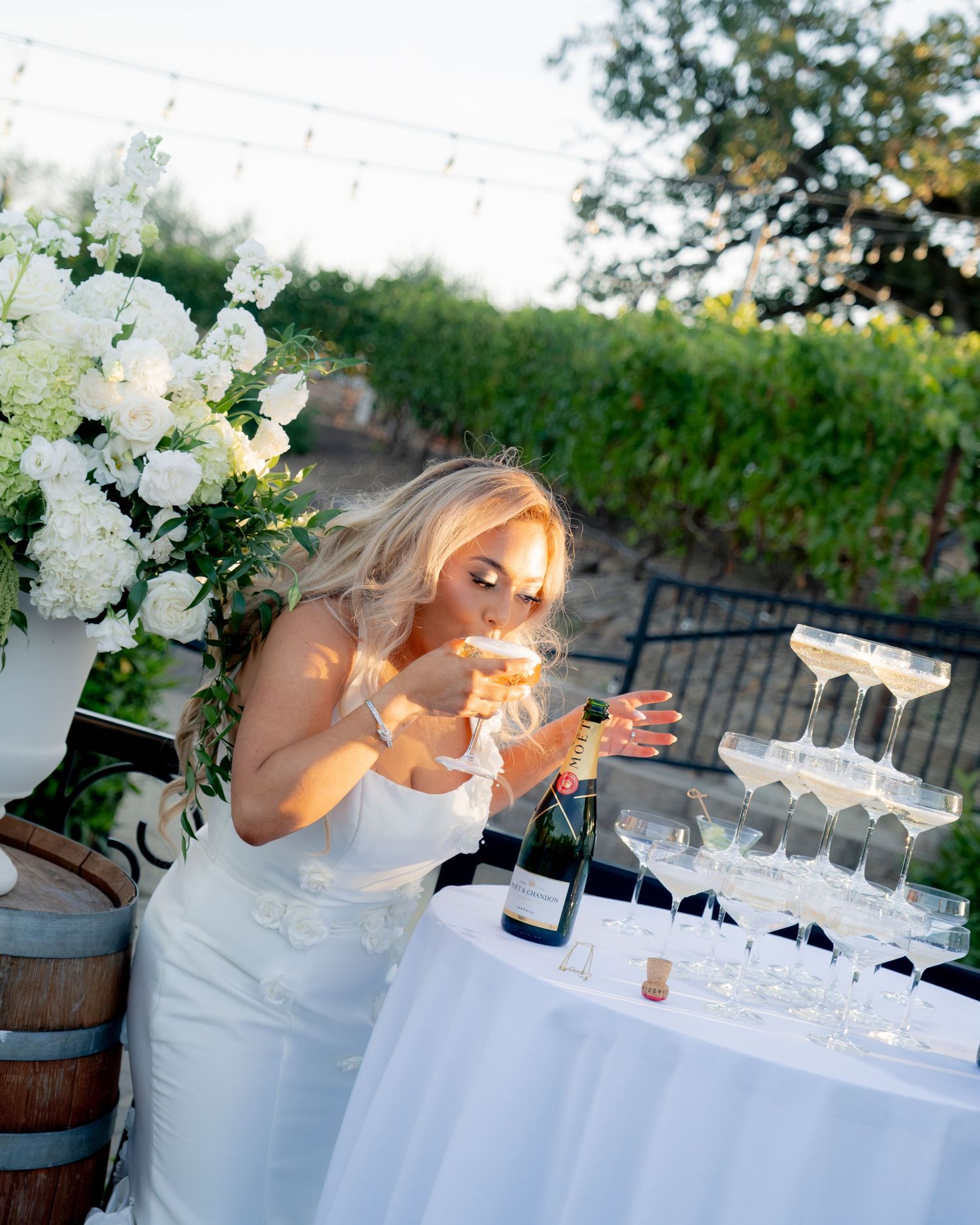 Blonde bride in white gown leans over champagne tower surrounded by white floral arrangements at outdoor reception.