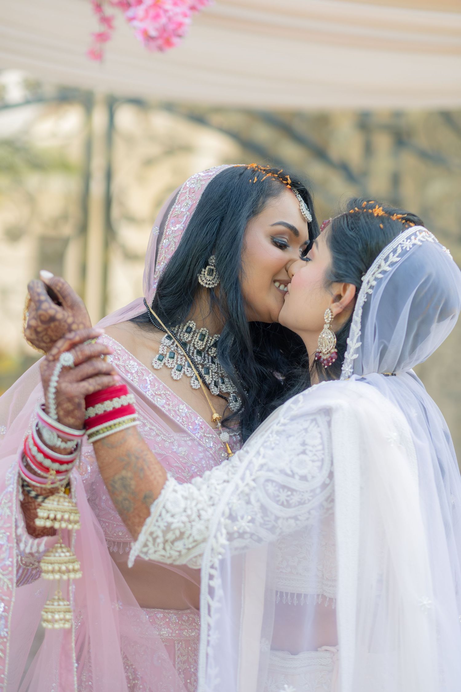 Two brides in traditional Indian bridal attire share a kiss during their wedding ceremony.