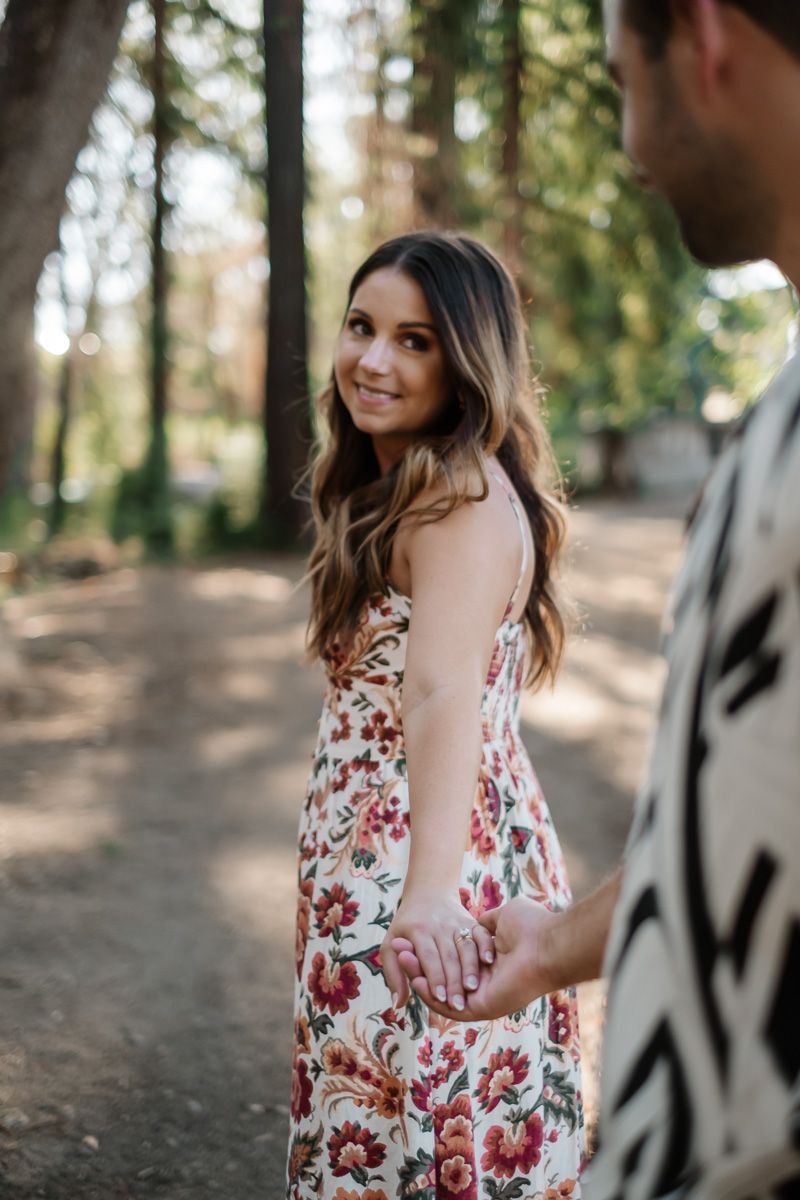 Young woman in a floral dress smiling over her shoulder while holding hands with her partner outdoors.
