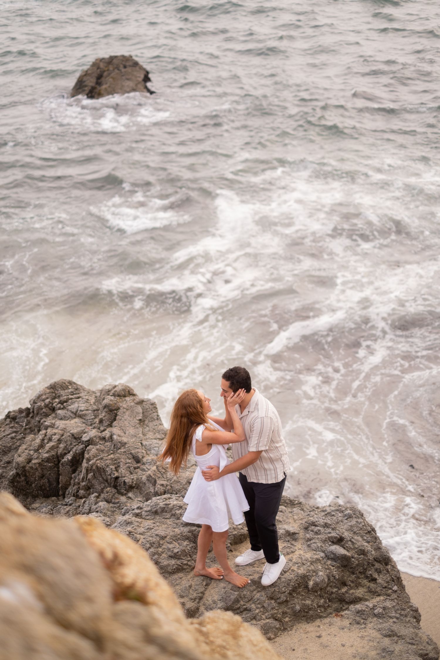 Couple embracing romantically on coastal rocks above crashing ocean waves, woman in flowing white dress.
